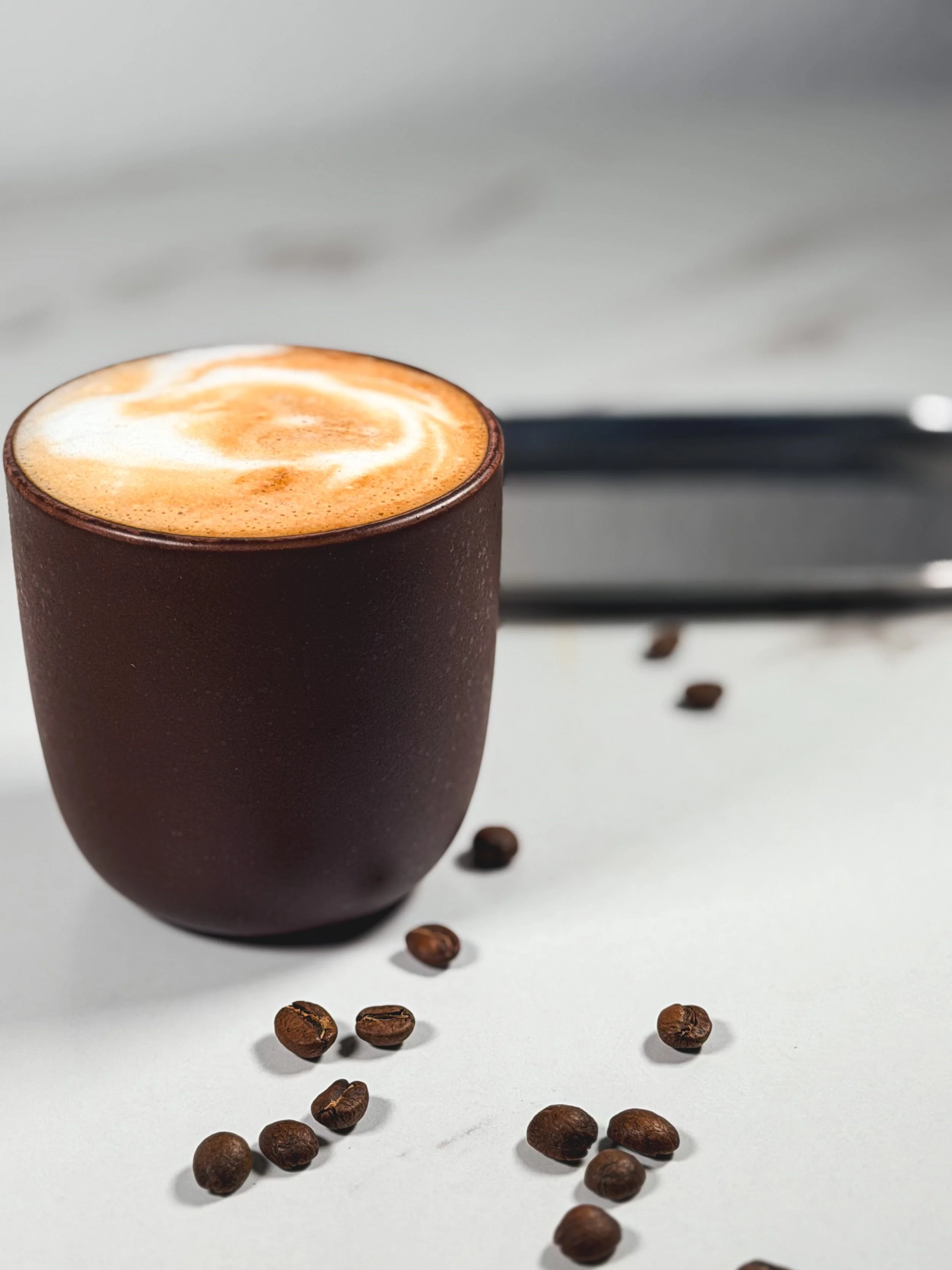A brown ceramic mug filled with frothy coffee or latte on a white surface, with coffee beans scattered around it.