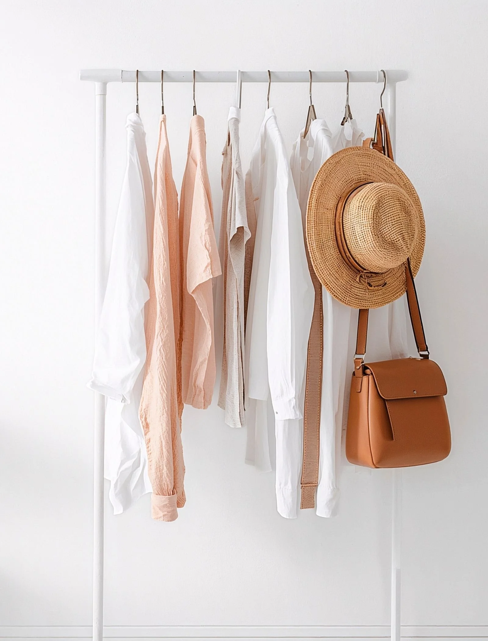 Clothing rack with white and light peach clothing, a straw hat, a brown purse, and wooden hangers, set against a plain white wall.