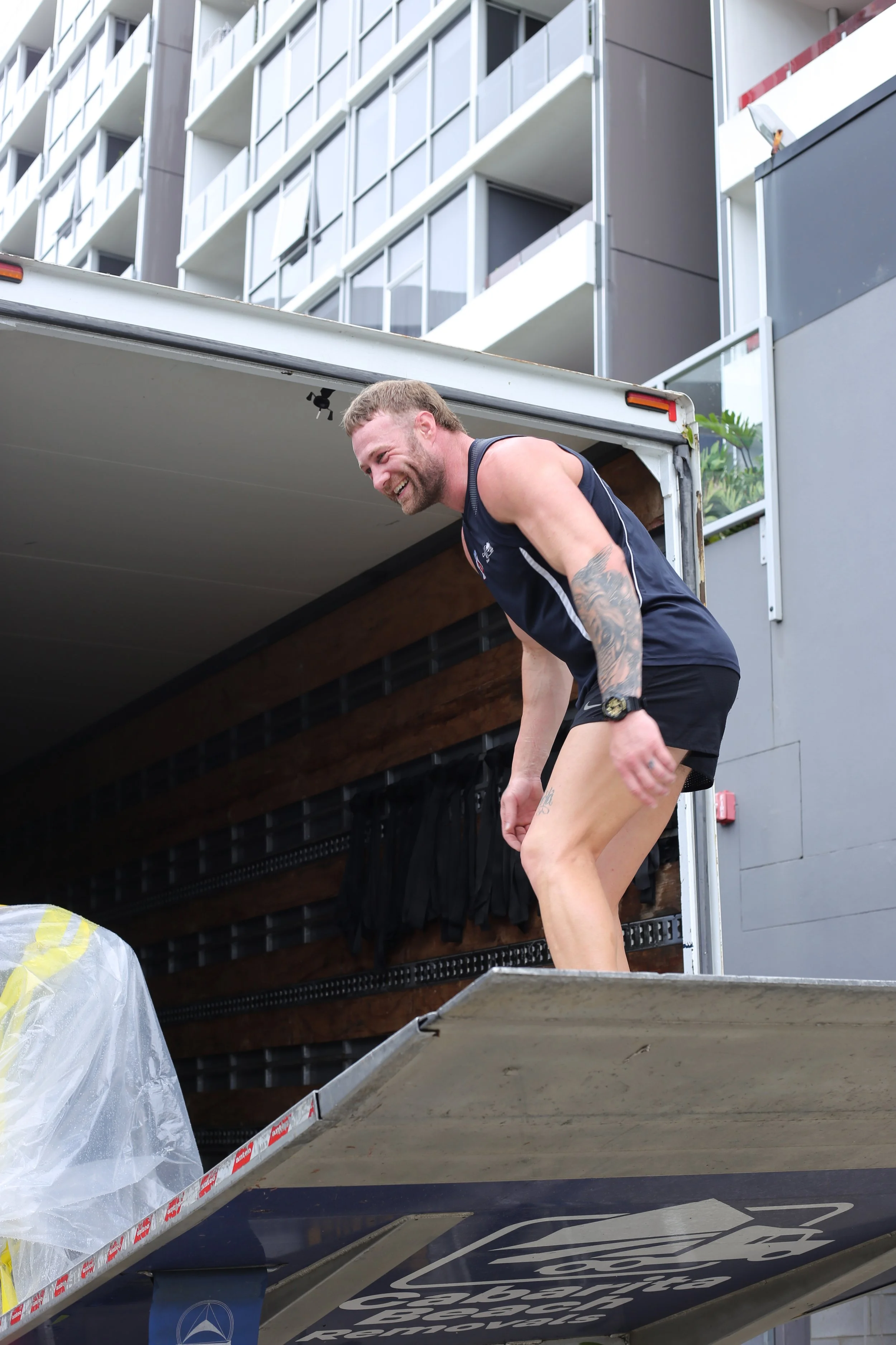 Tattooed man in athletic clothing unloading a moving truck outside a modern apartment building.