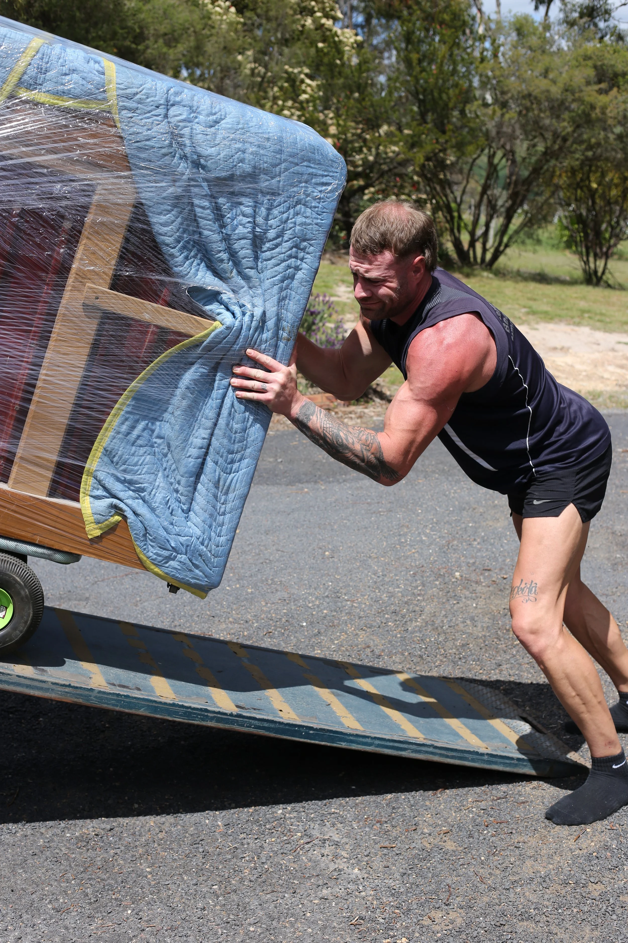 Man securing a large blue padded object wrapped in plastic on a wheeled platform outdoors