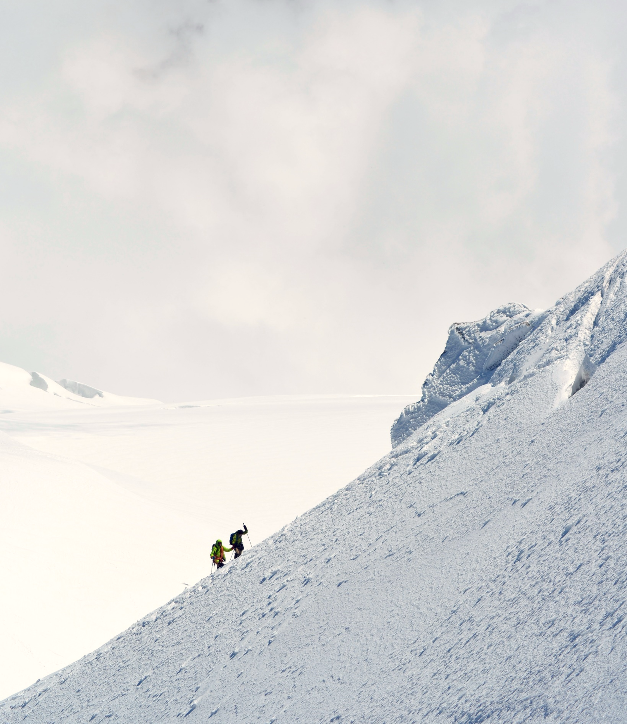 Two climbers in colorful gear ascending a snow-covered mountain slope with a backdrop of snow-capped peaks and a pale sky.