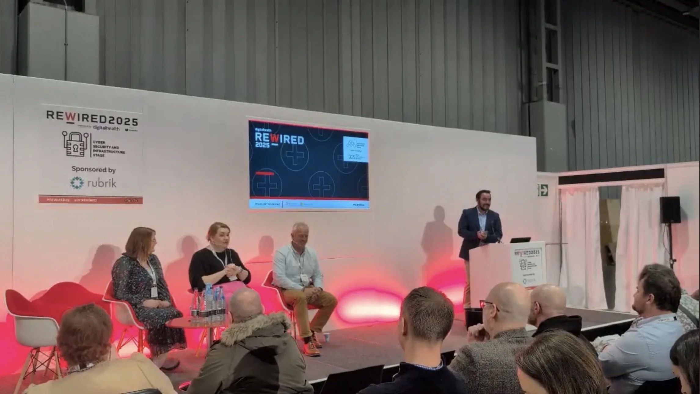Panel discussion at REWIRED 2025 event, with four people seated on stage and one person speaking at a podium, audience watching in a large indoor venue.