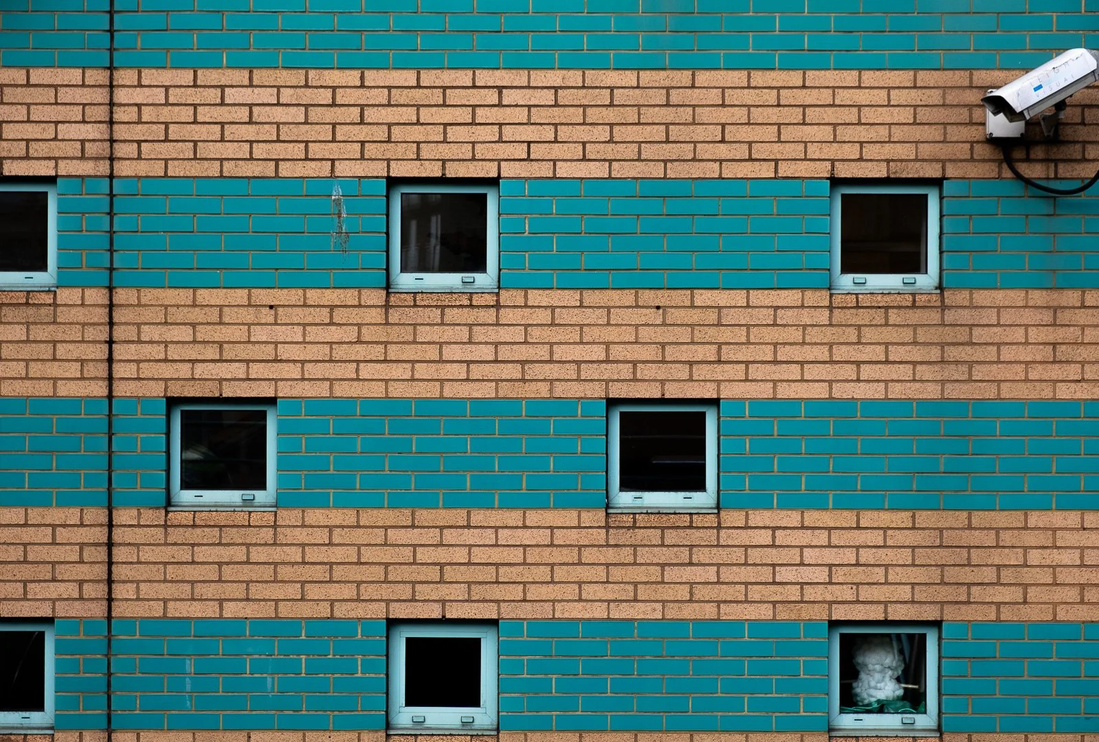 Close-up view of a multistory building's exterior wall made of beige and turquoise bricks, with several small square windows, some with reflections and one with cotton or insulation material visible inside. A security camera is mounted on the upper r