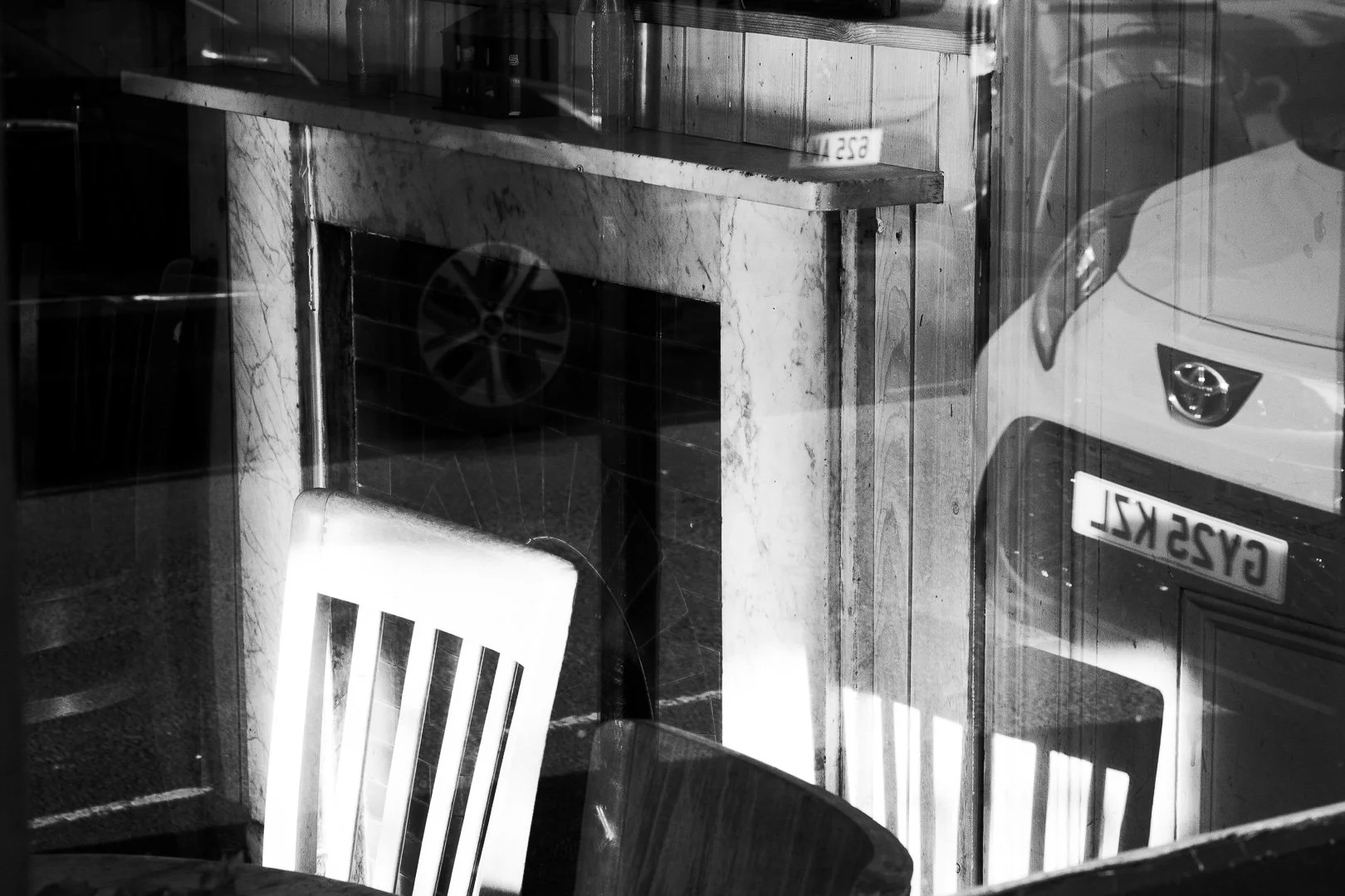 Black and white photo of a cafe interior and a reflection of a white car with a visible license plate reading 'G52  VKL' in a window.