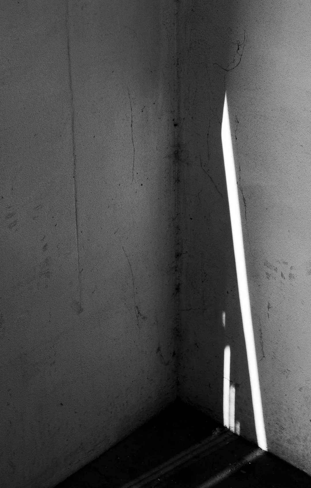 A corner of a room with cracked and dirty white walls and a narrow vertical opening with a strip of bright sunlight casting a shadow.