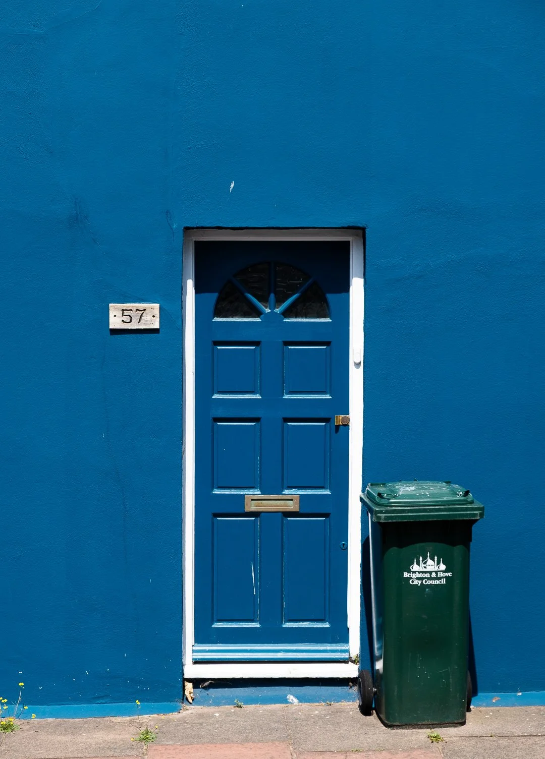 Bright blue front door with a semi-circular window at the top, set into a matching blue wall. To the left, a house number plaque reads '57'. To the right, a green trash bin marked 'Brighton & Hove City Council' is positioned on the sidewalk.