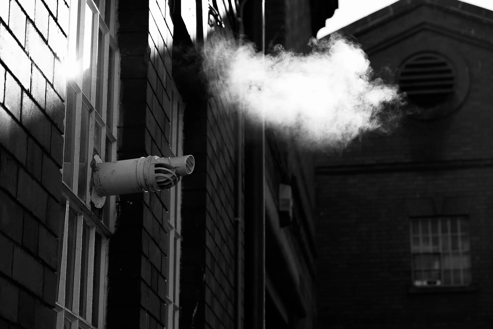 A black and white photo of a pipe emitting white smoke or vapor from the side of a brick building, with the sun shining on the brick wall.
