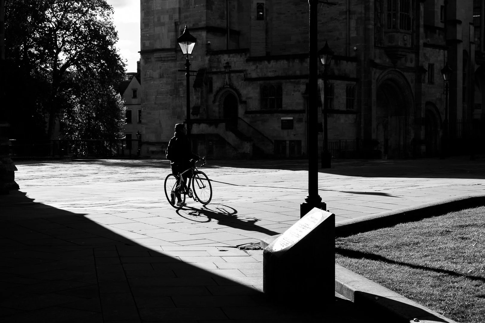 A person on a bicycle, shadow cast on the sidewalk, with an old stone building and street lamps in the background, in a black and white photo.
