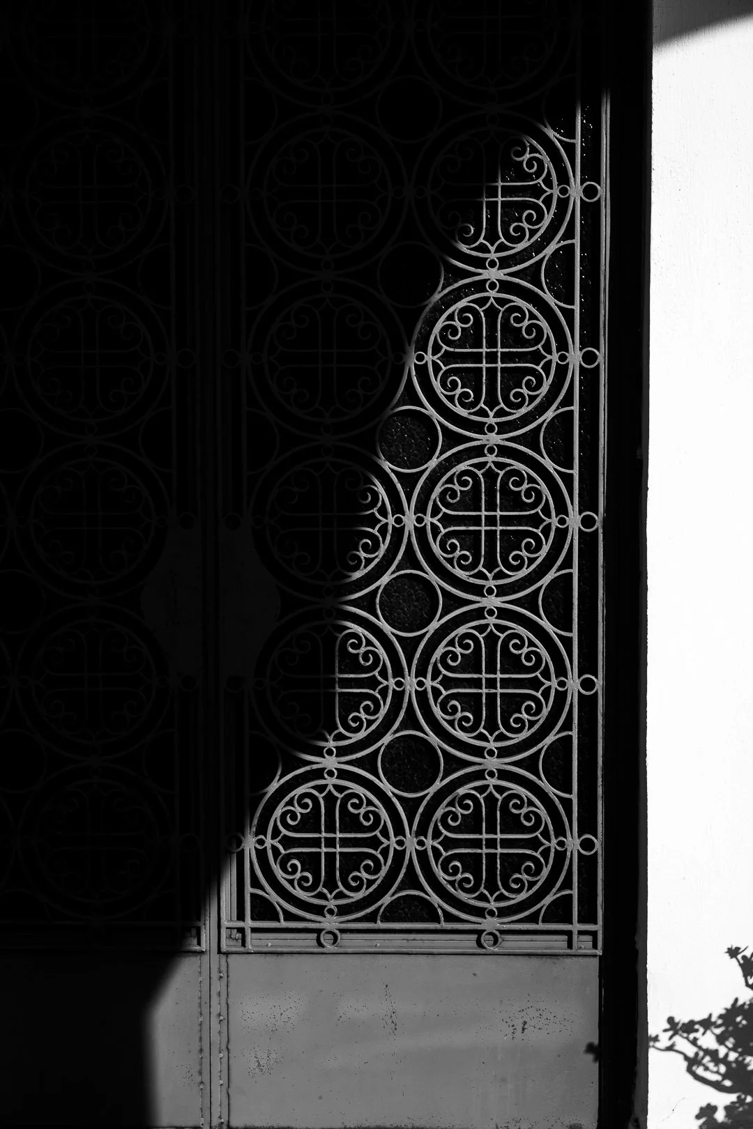 Black wrought iron gate with circular and swirl patterns, partially illuminated, next to a white wall, with shadows cast on the left side.