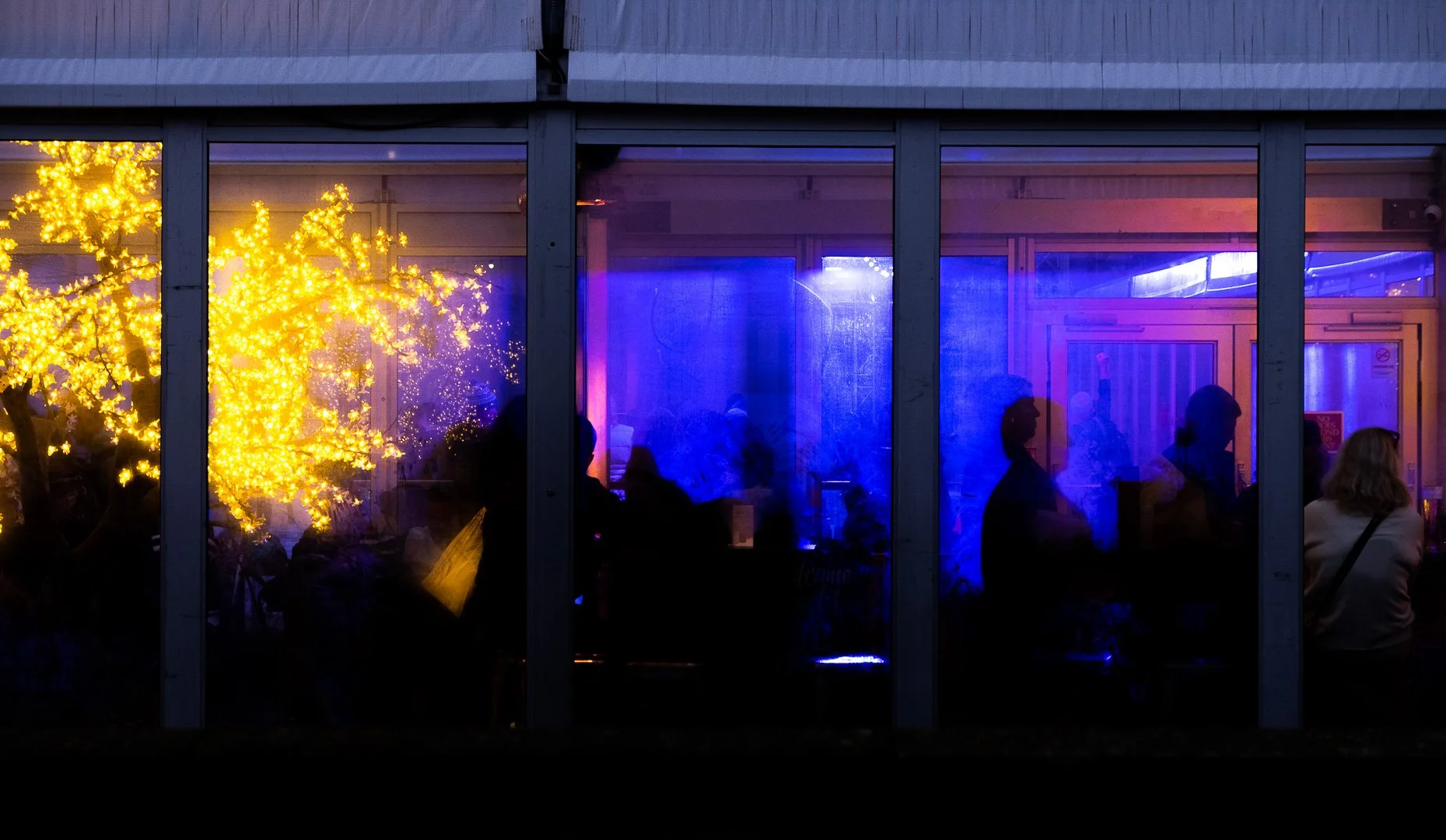 People sitting inside a building at night, visible through large glass windows. The interior is illuminated with yellow and blue decorative lighting, including a yellow-lit artificial tree to the left.