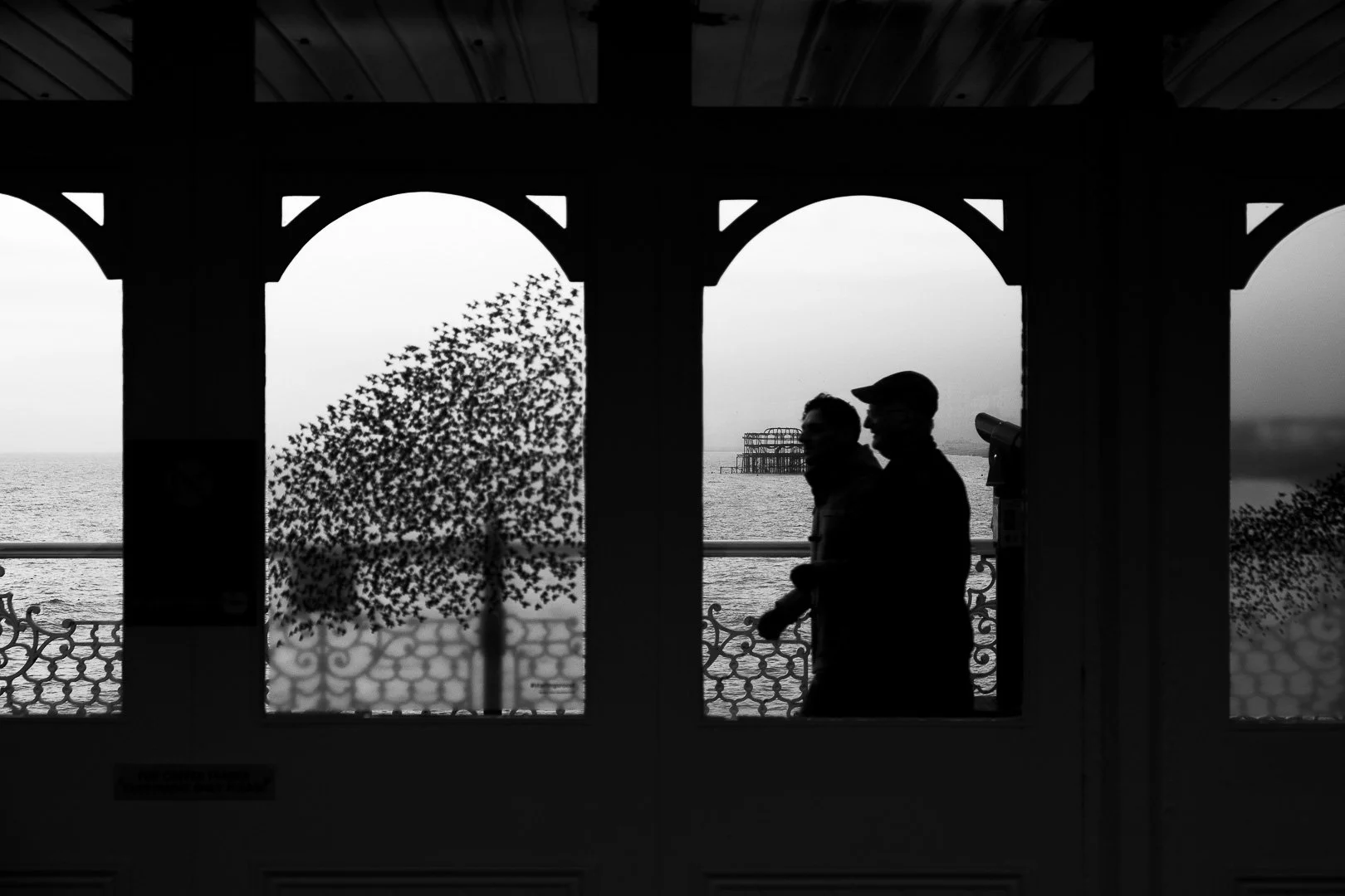 Silhouettes of two people walking along a seaside promenade during daytime, seen through arched windows with an ocean and a distant pier in the background.
