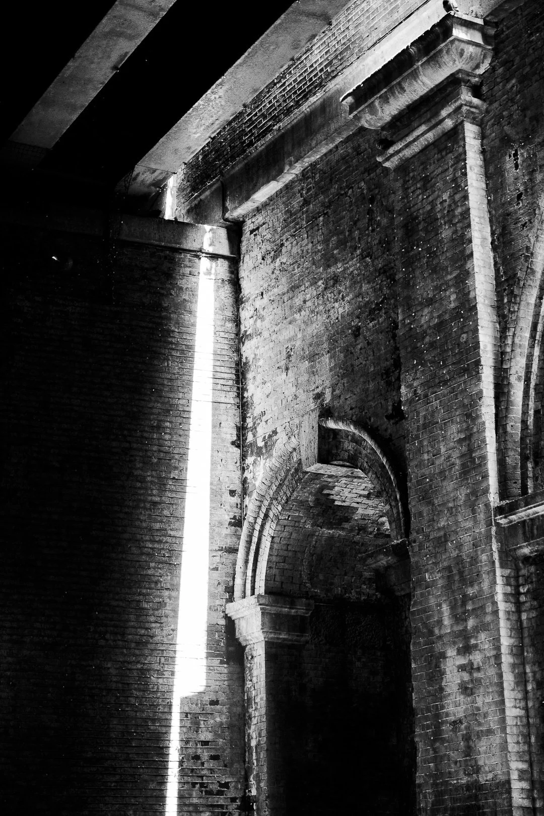 Black and white photo of an old brick building's interior, highlighting a large archway and a vertical beam of light coming through a small opening.