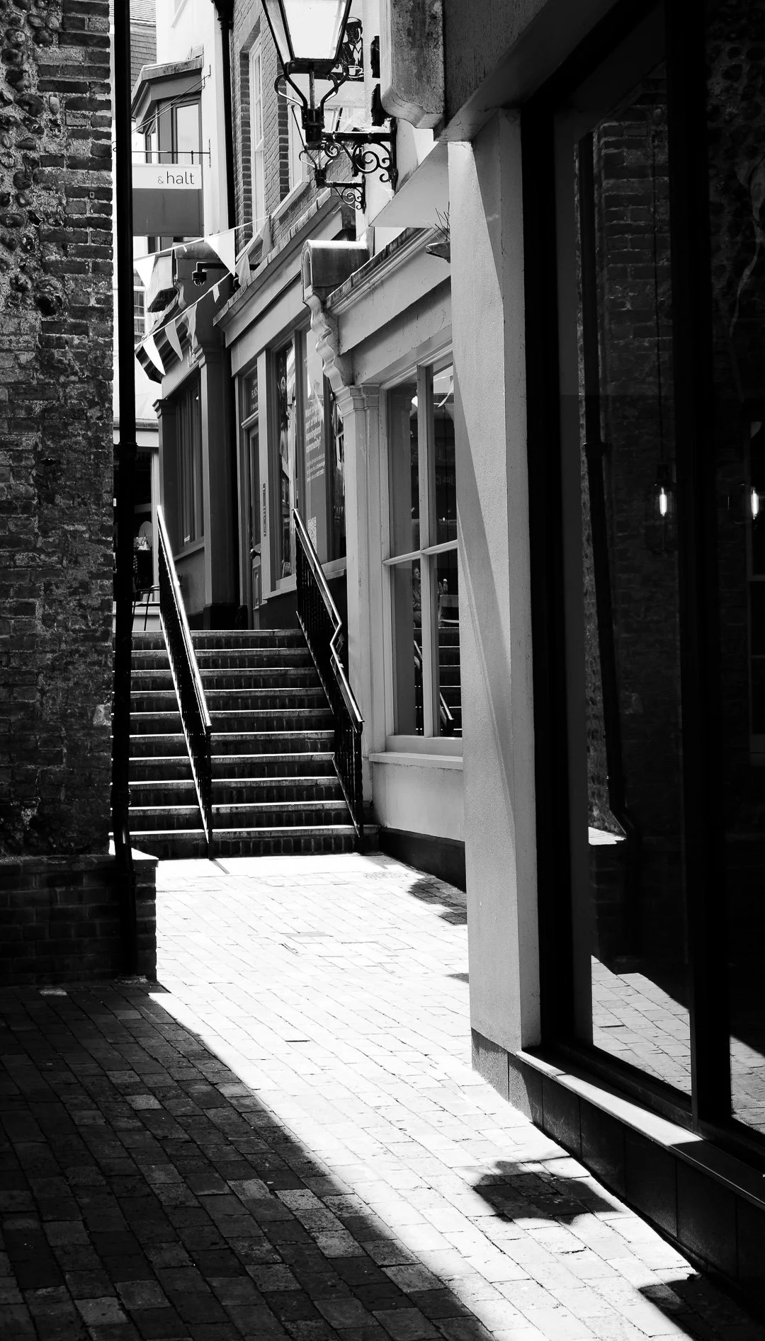 Black and white photo of a narrow alleyway with stairs leading up to a building with large windows. A sign with the word 'halt' is visible hanging from a building on the left. Shadows and sunlight create contrast on the brick and wall surfaces.