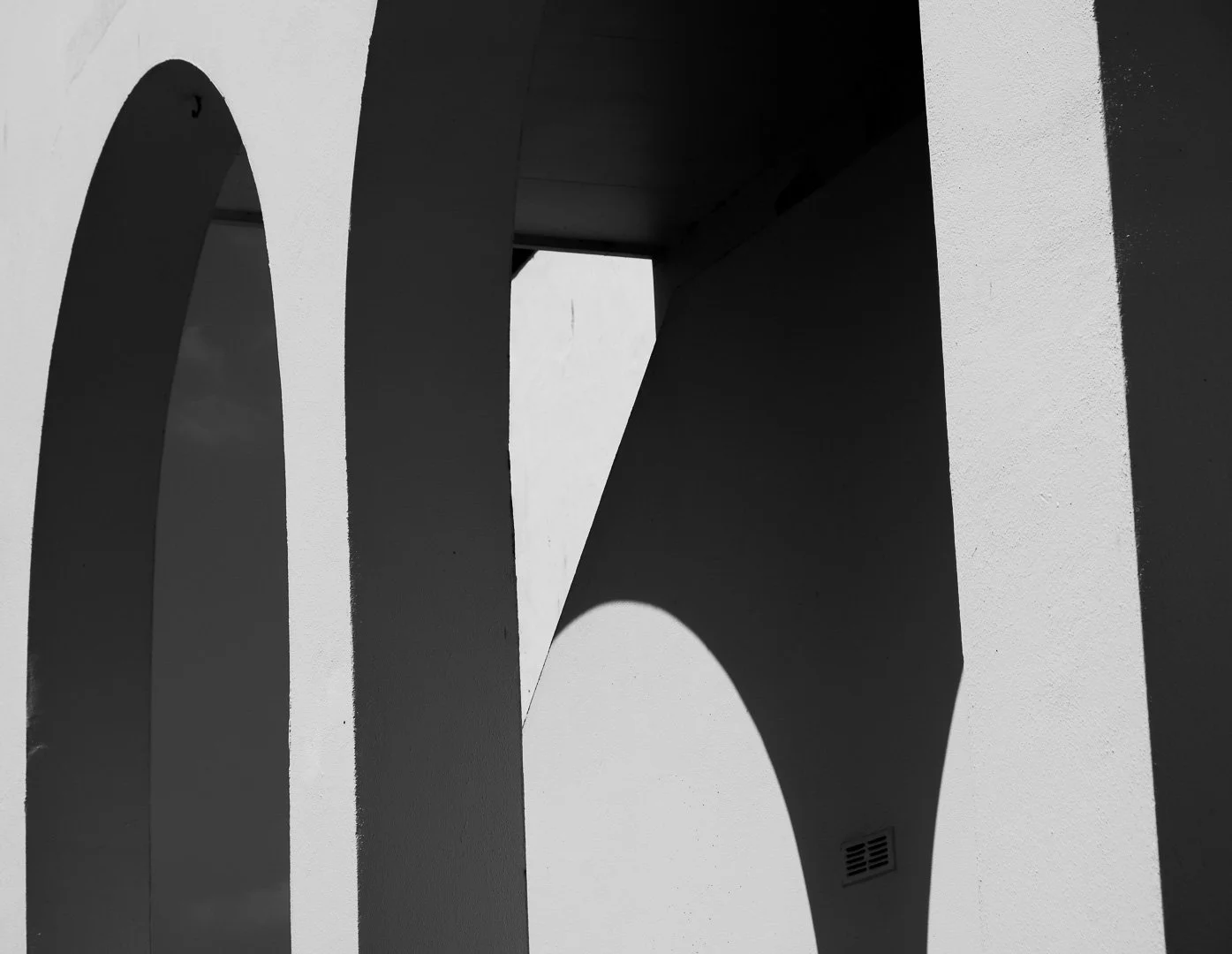Black and white photograph of geometric architectural elements with shadows creating abstract shapes on a white wall, including arches and angular forms.