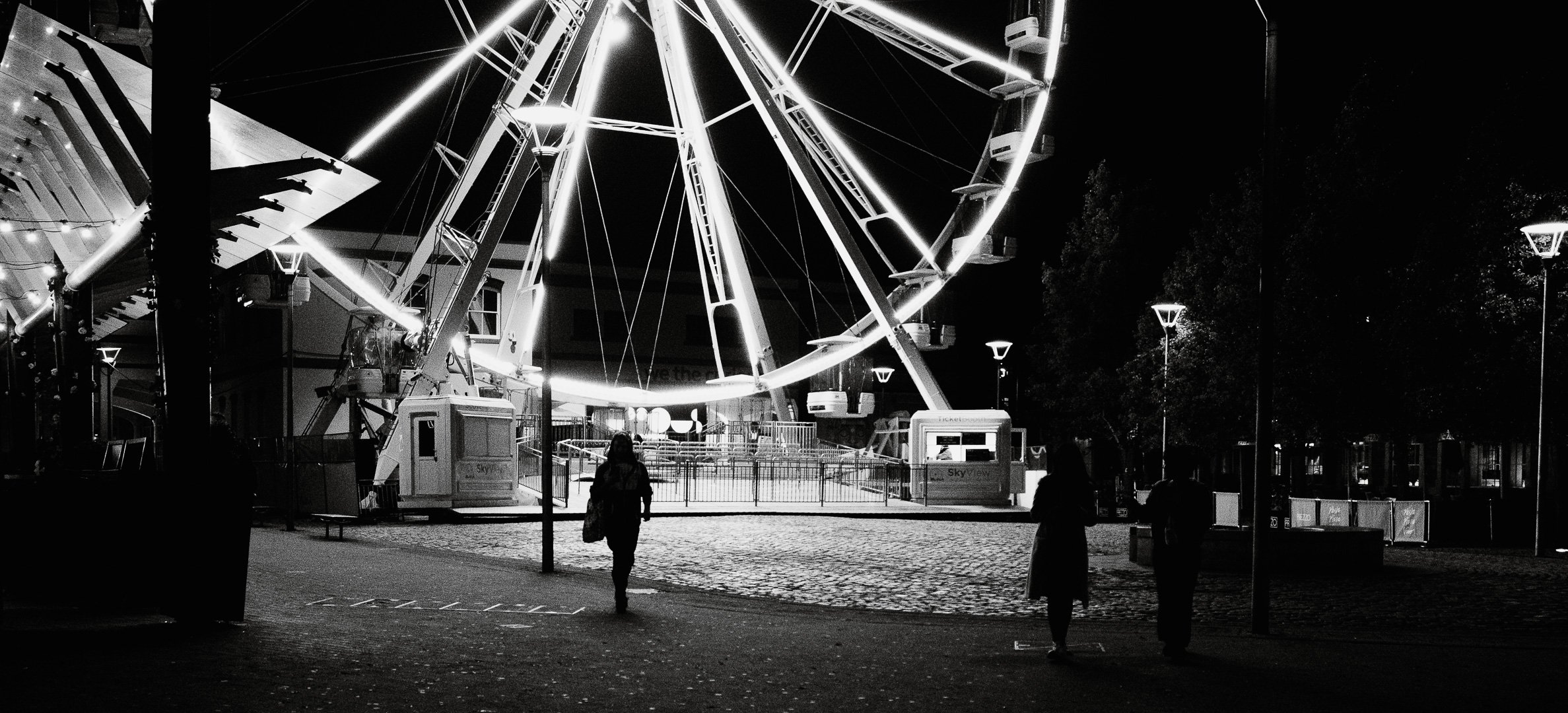 Nighttime scene of a carnival with a large illuminated Ferris wheel, empty rides, and a few silhouetted people walking or standing around.
