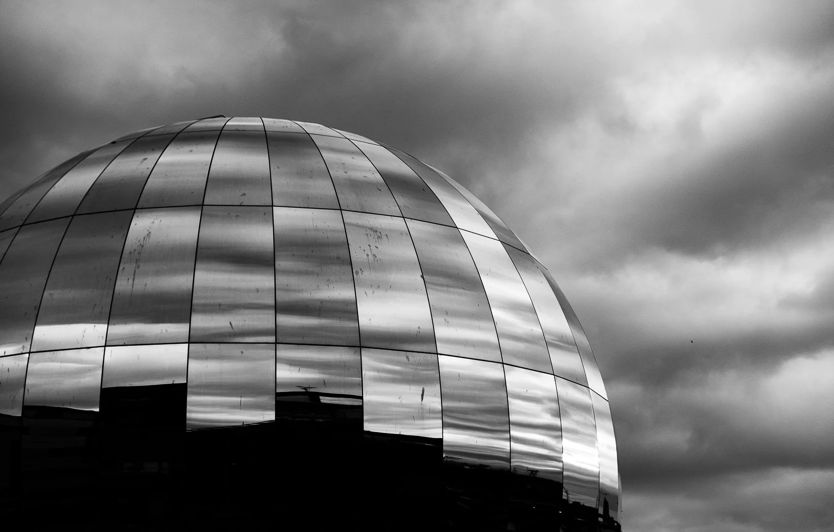 Black and white photo of a modern, dome-shaped building with reflective metallic panels and a cloudy sky.