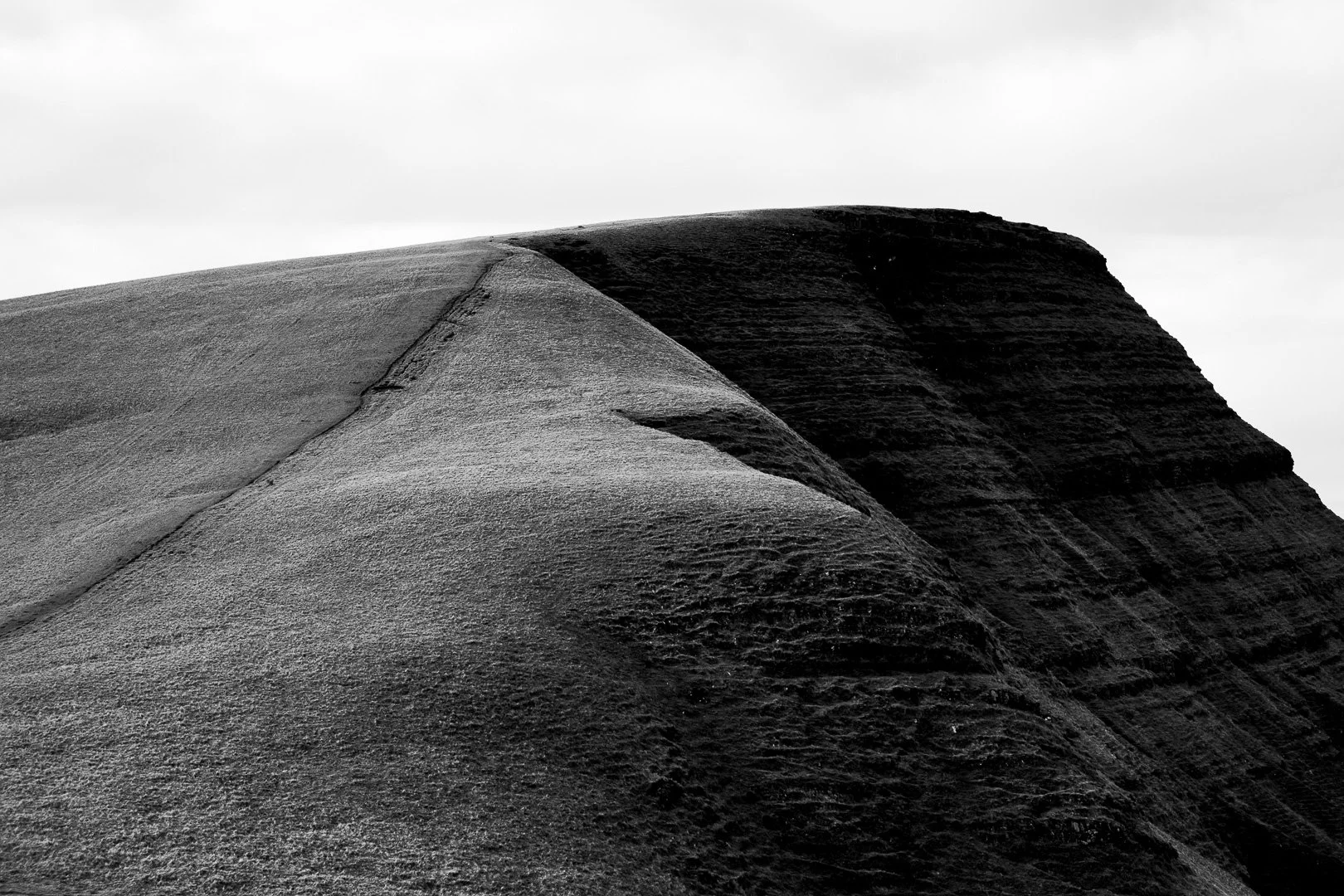 Black and white photograph of rolling grassy hills with textured surfaces and gentle slopes under an overcast sky.