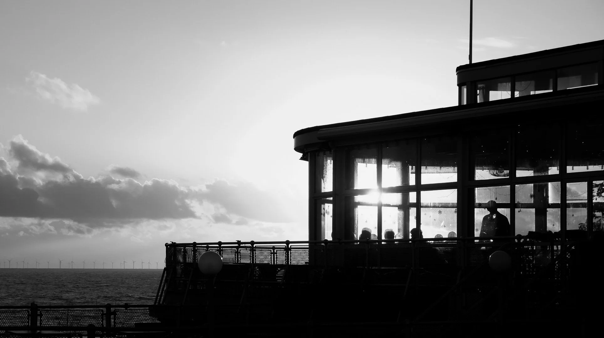 Black and white photo of a circular glass building on the ocean with wind turbines on the horizon, sunset or sunrise backdrop with clouds.
