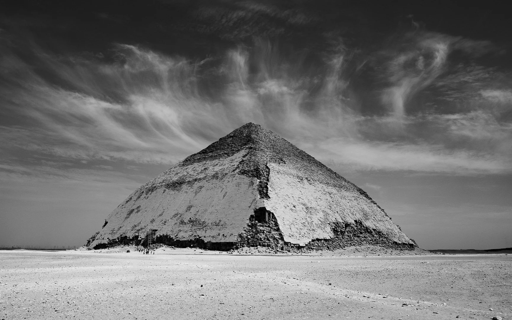Black and white photo of a large pyramid-shaped structure with steps and ruins at the base, set in a barren, flat landscape under a partly cloudy sky.