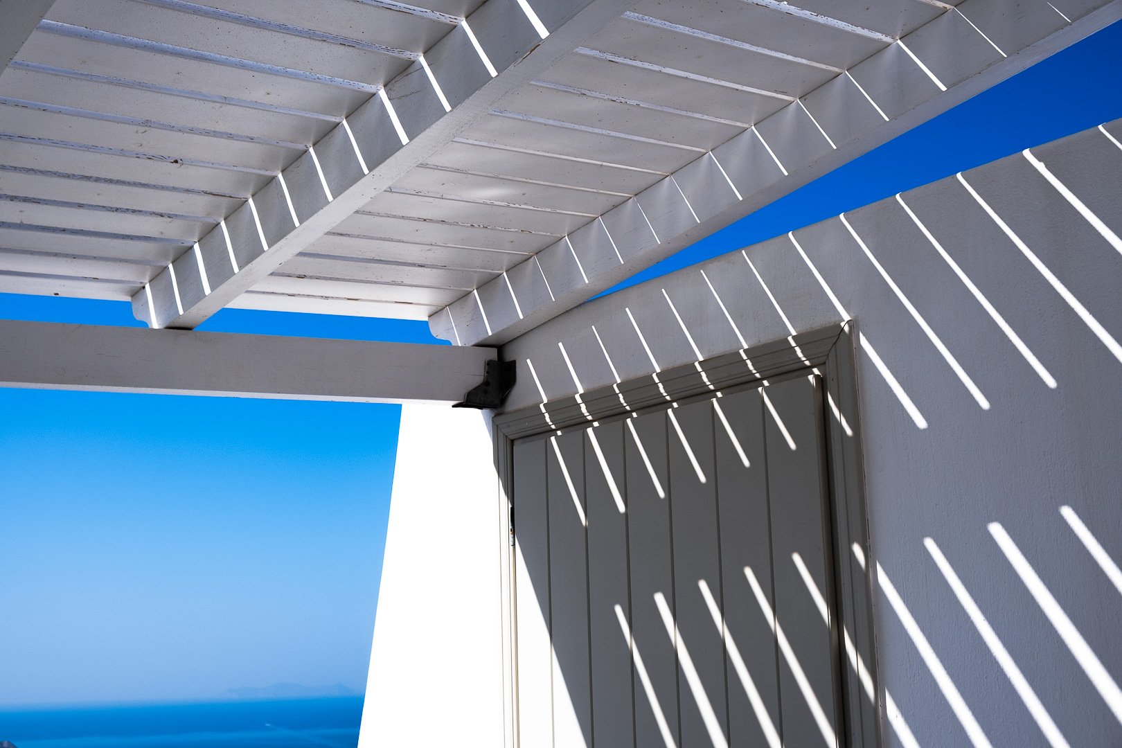 An outdoor space with a white slatted pergola casting striped shadows on a white wall and door, against a clear blue sky.