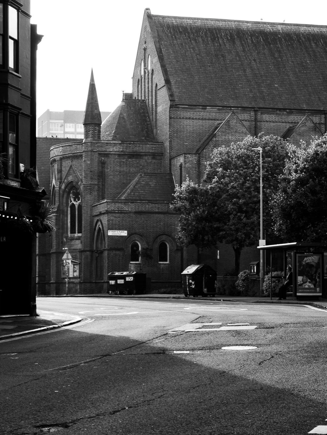 Historic brick church with pointed steeples on a city street, with trees, a bus stop, and garbage bins in the foreground, and modern buildings in the background.