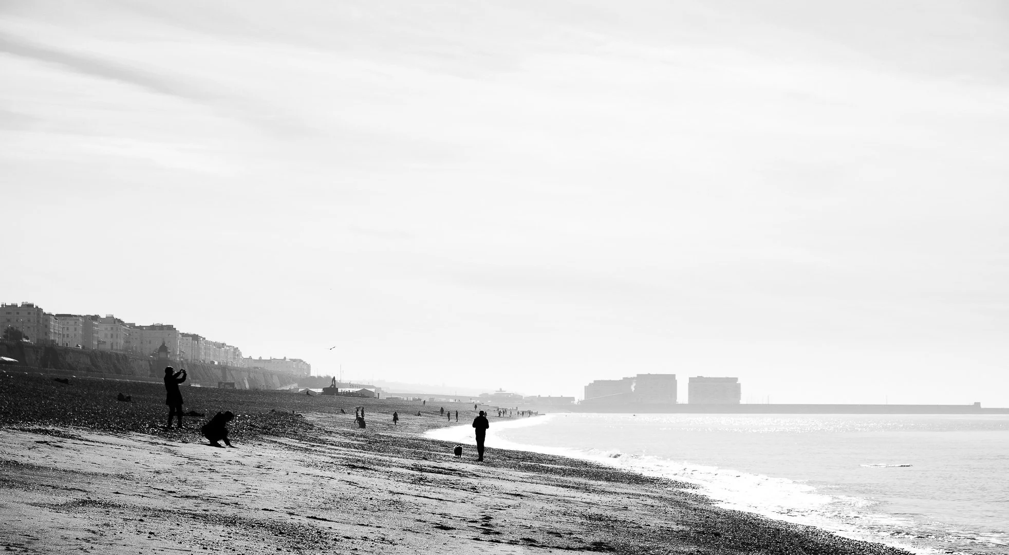 Black and white photograph of people walking and relaxing on a beach with a city skyline in the background.