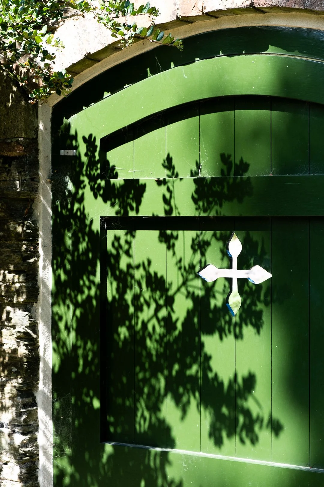Green door with a white cross-shaped decoration, shadows of leaves cast on the door, stone and brick wall on the side, foliage at the top.