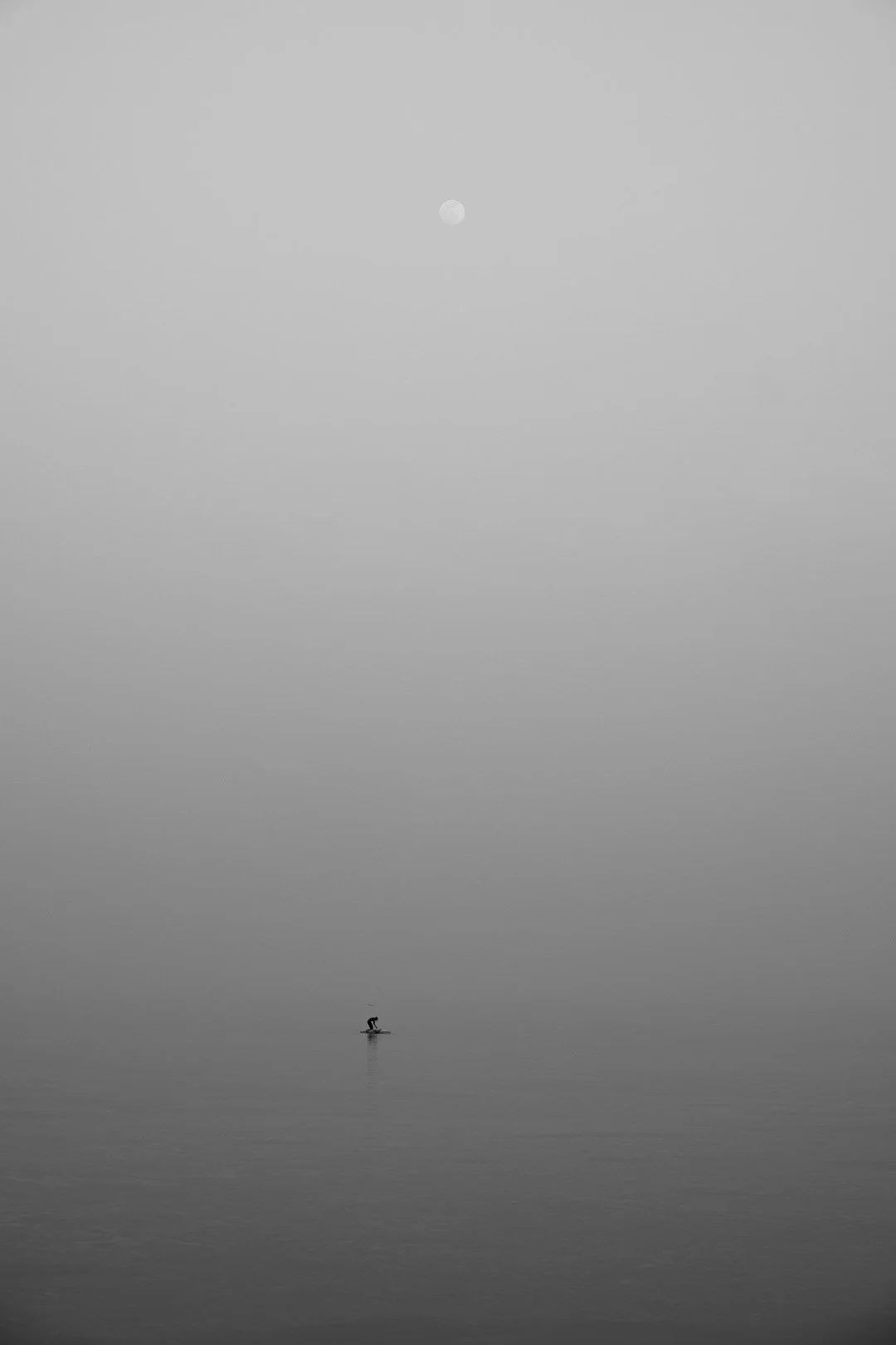A person paddleboarding on calm water under a cloudy sky with the moon visible in the distance.