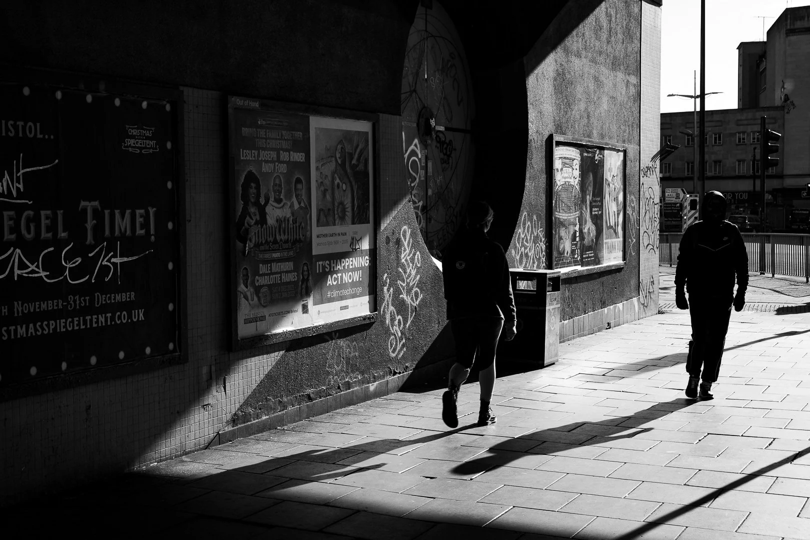 Two people walk along a sidewalk under a bridge or tunnel in sunlight, with posters on the wall and graffiti, and city buildings and traffic lights in the background.