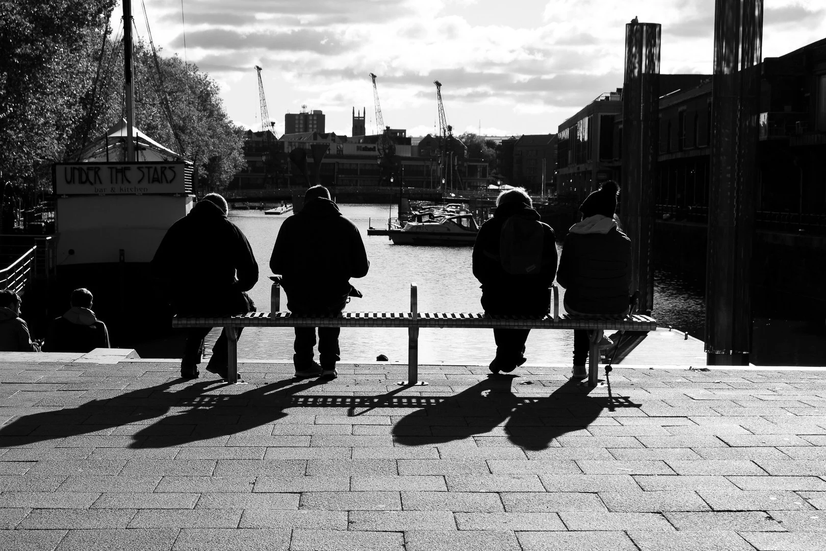 Four people sitting on a bench by a waterfront, with three children sitting nearby, and sailboats and cranes in the background, in black and white.