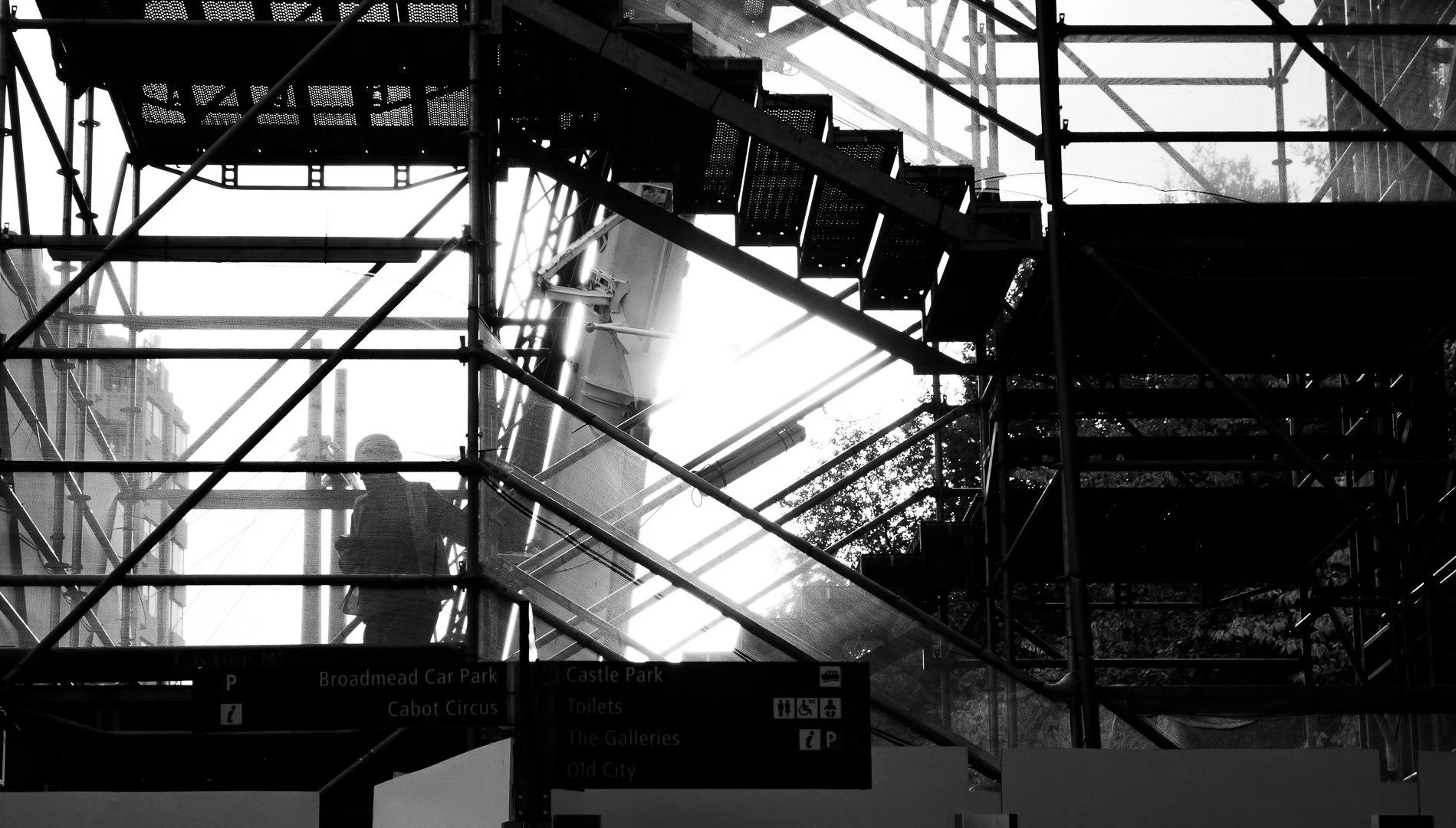 A person wearing a hard hat and covering their face with a mask, standing on scaffolding at a construction site with metal framework and stairs, during daytime with bright light in the background.