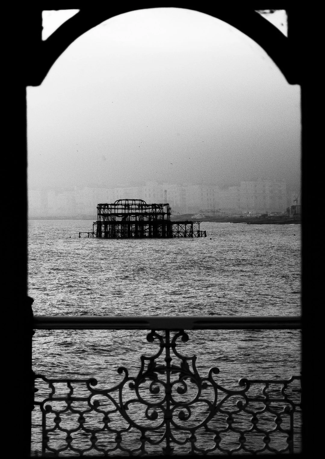 Silhouette of a bridge archway overlooking a body of water with a partially submerged, dilapidated pier or structure in the distance, foggy city skyline in the background, black and white photo.