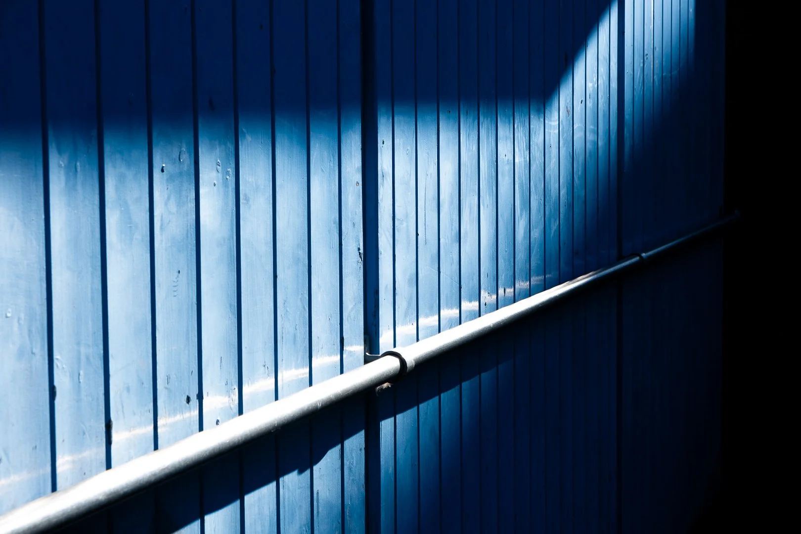 Close-up of a blue wooden wall with a silver metal pipe running horizontally across it, with sunlight creating a diagonal shadow on the wall.