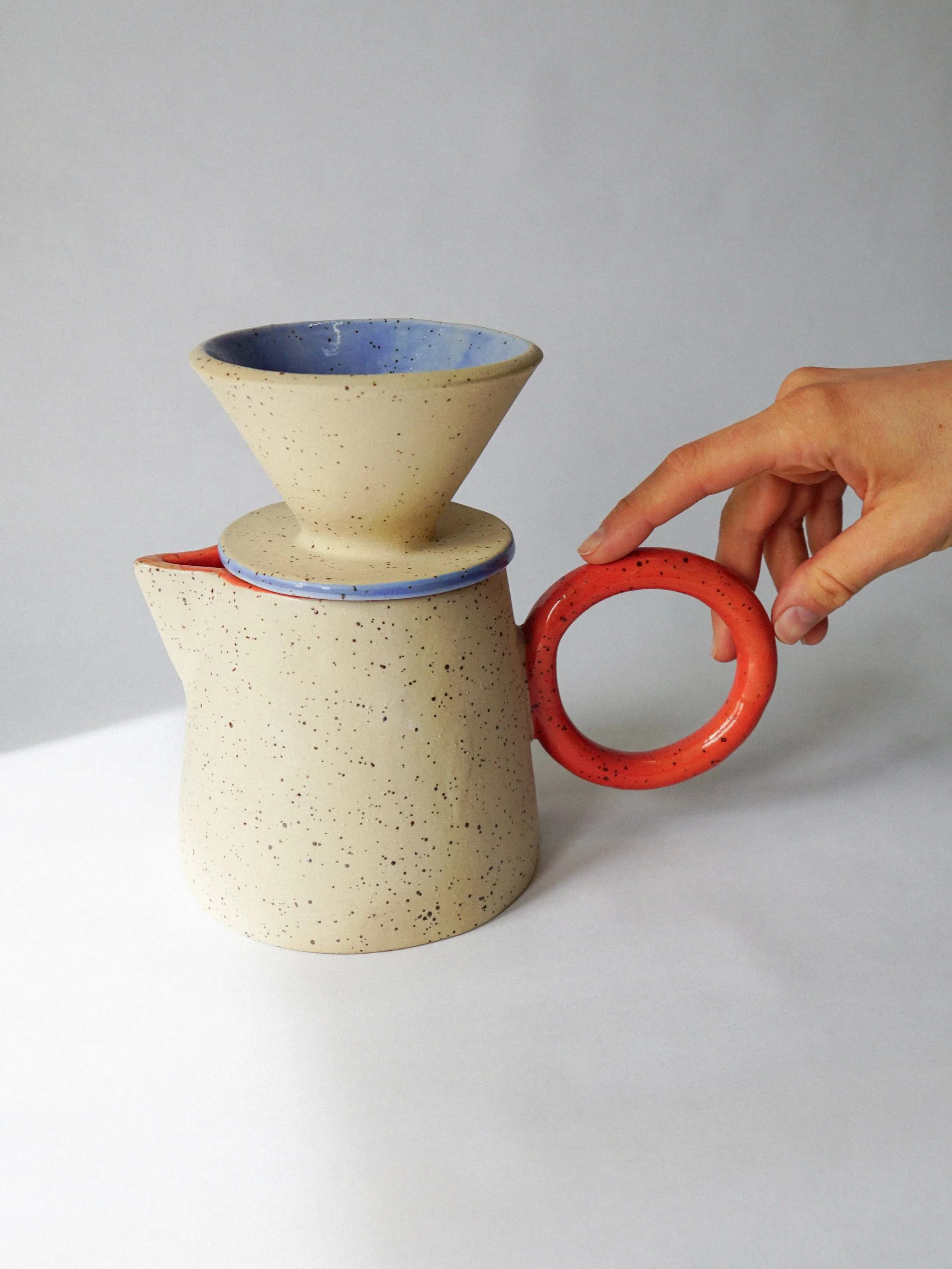 Hand placing a red-handled ceramic lid on a speckled stoneware teapot with a blue-rimmed lid.