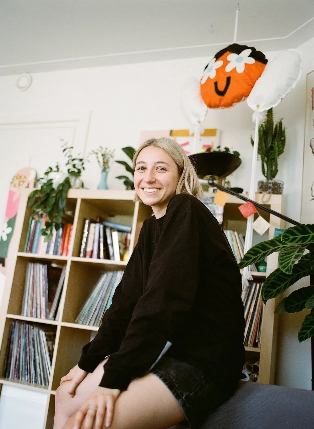 Smiling woman in a black outfit sitting in a plant-filled room with bookshelves, records, and a floral orange balloon.