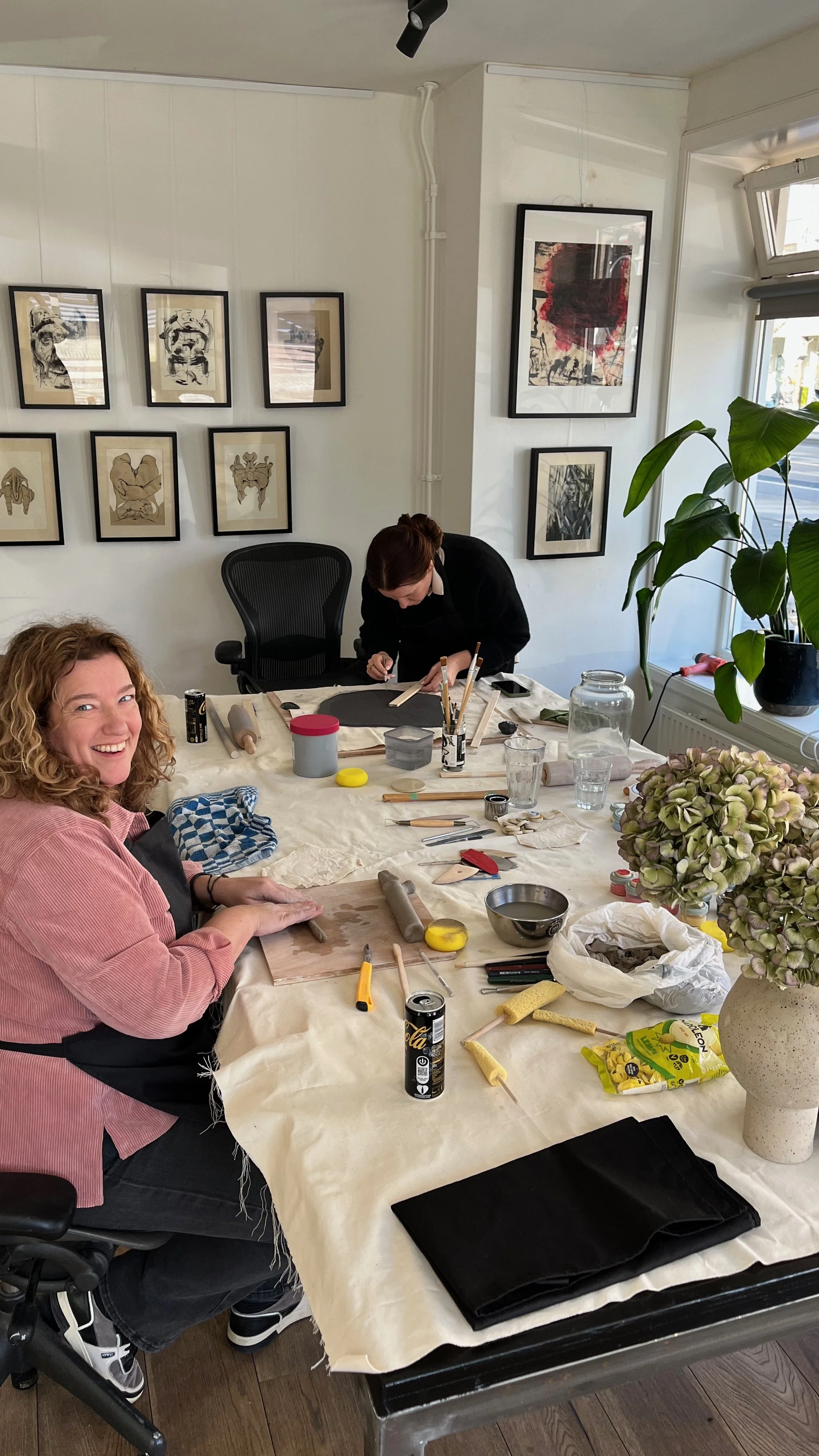 Two people work on a craft project at a table covered with art supplies in a bright studio displaying framed artwork and houseplants.