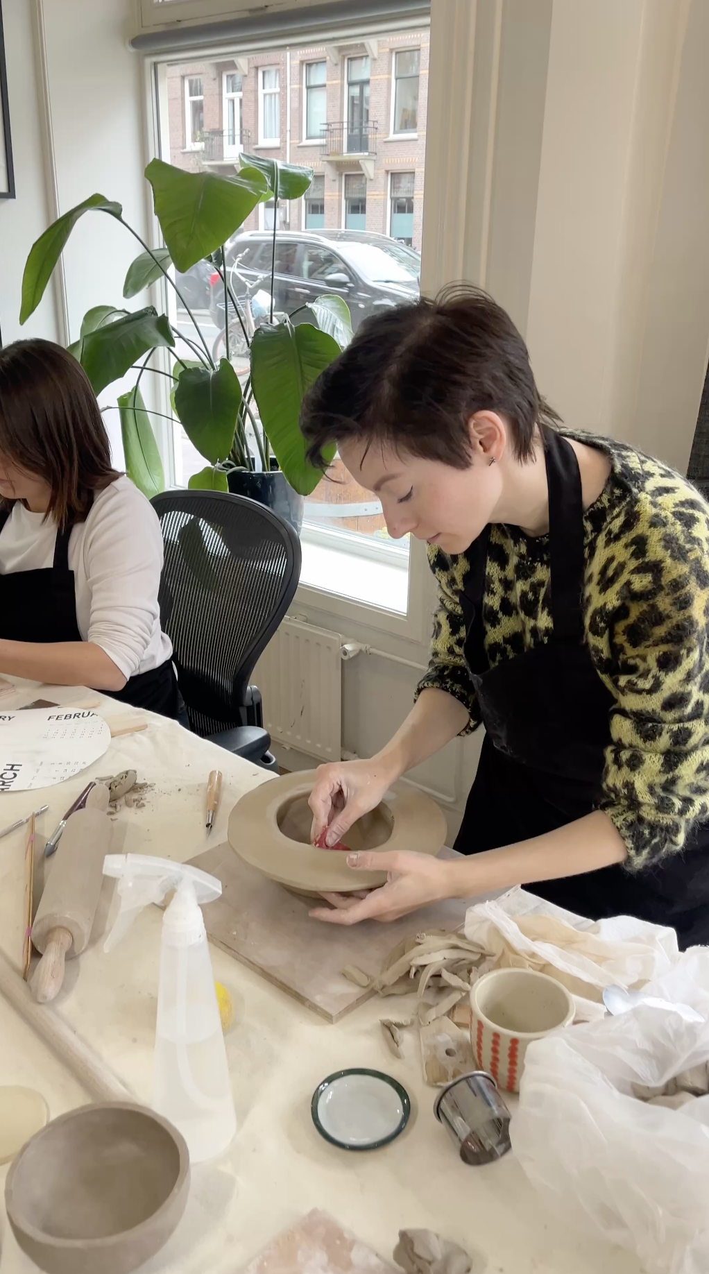 Two people shaping clay pottery at a ceramics studio table beside a window