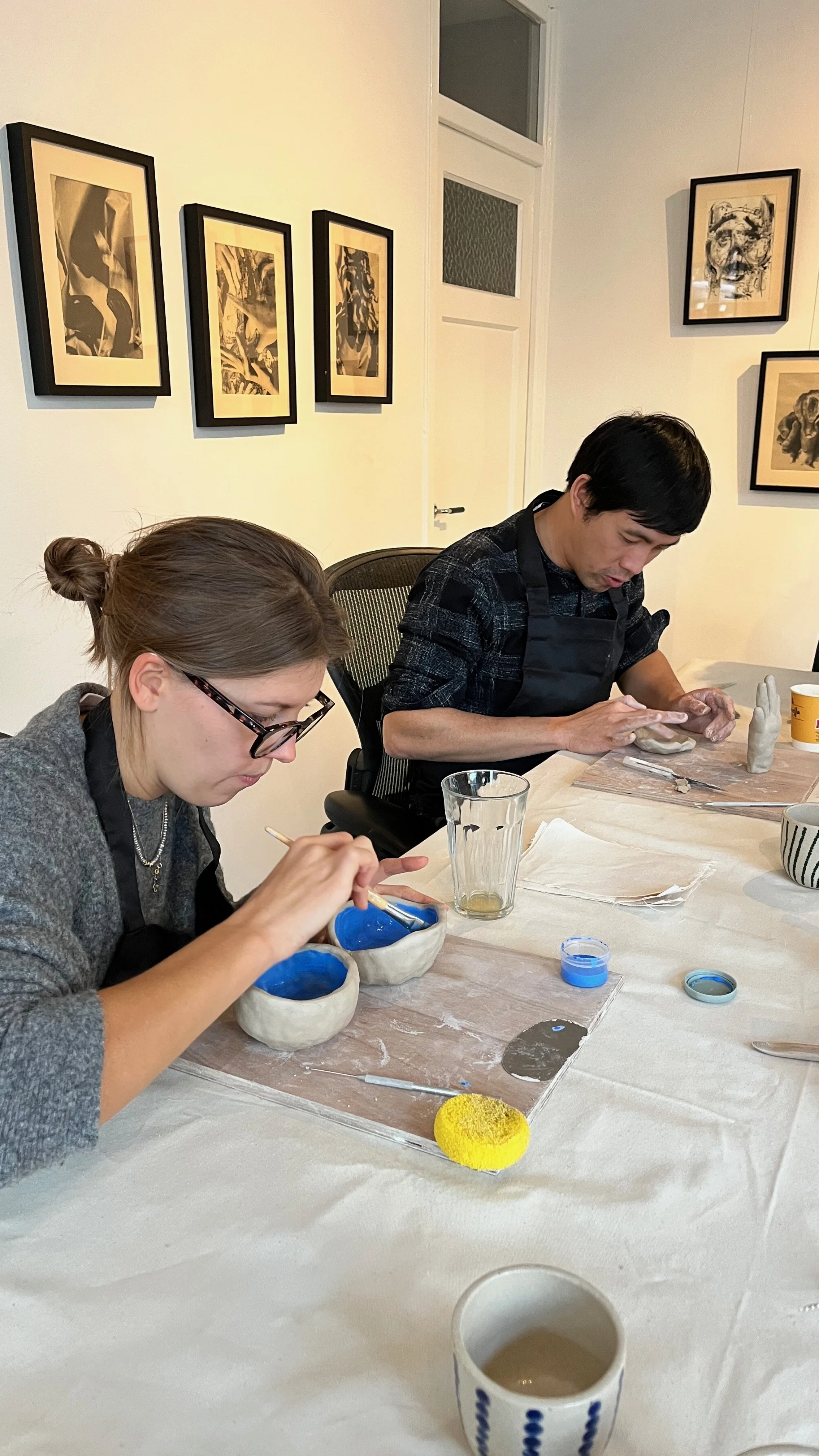 Two people shape clay at a pottery workshop table with blue glazes, tools, and framed artwork on the wall.