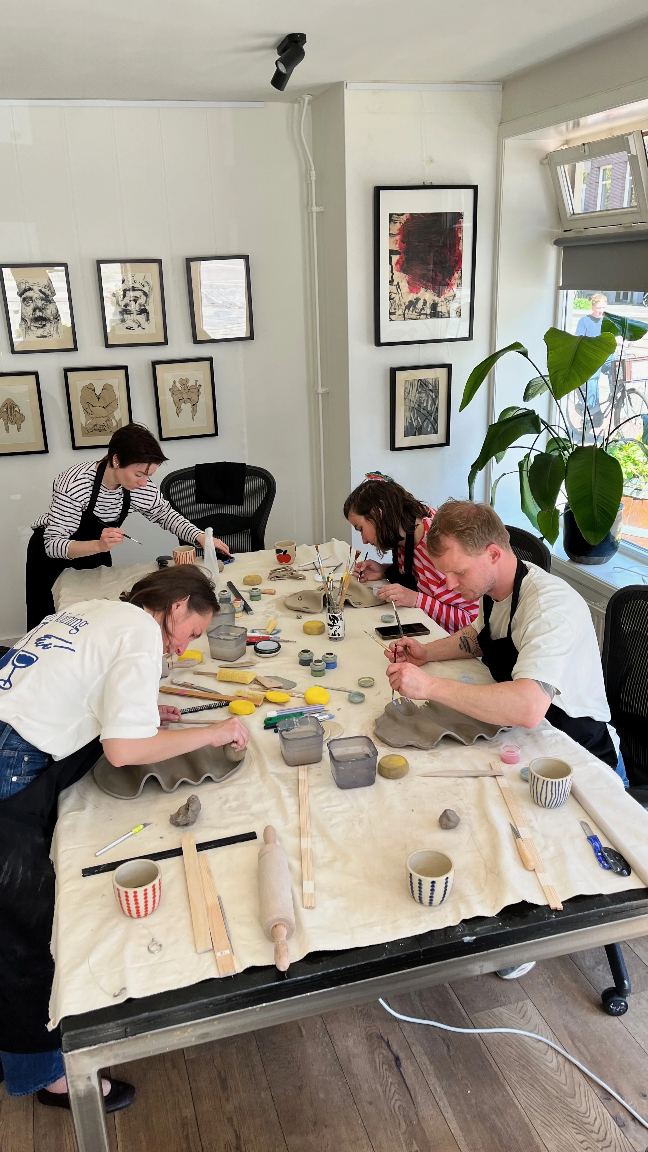 People painting handmade ceramic pieces during a pottery workshop at a table surrounded by framed artwork.