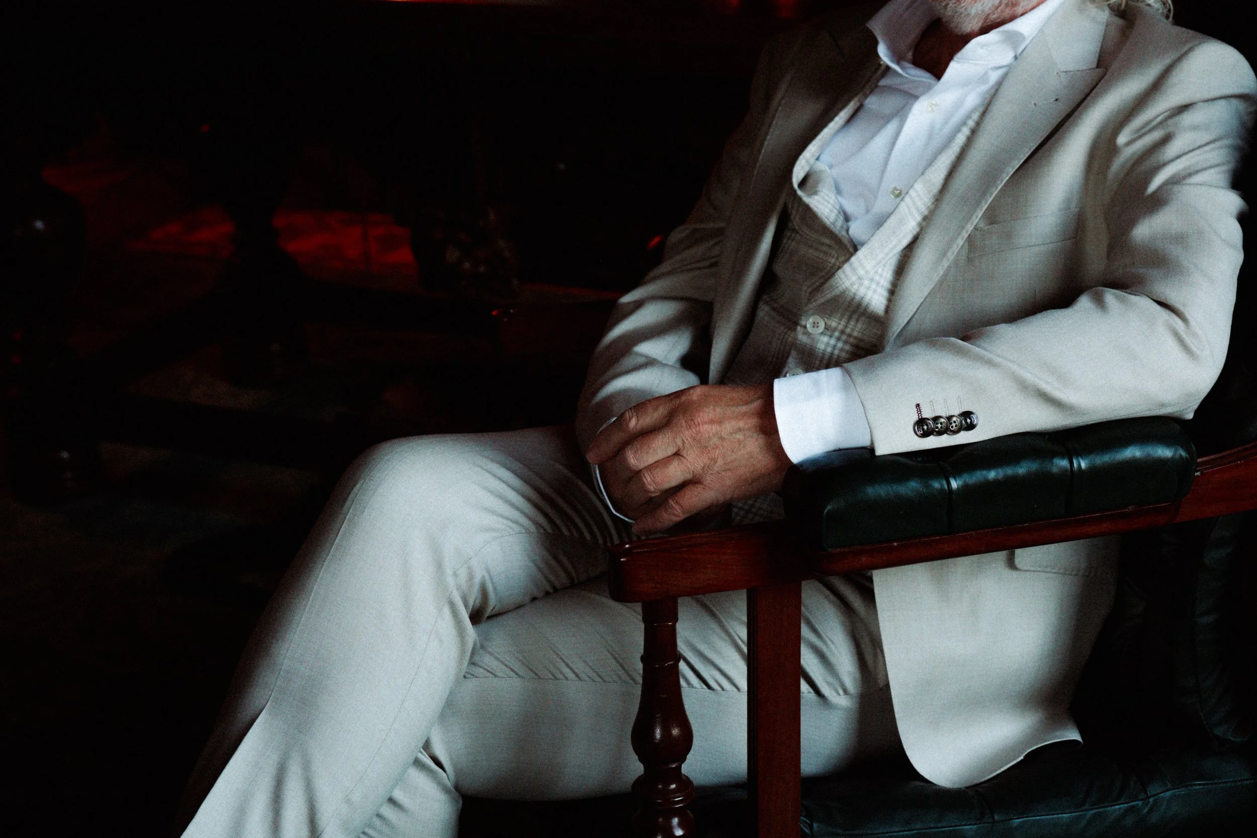 Man in een witte pak zit op een stoel met een houten armleuning, zijn armen gekruist, in een donkere omgeving. bruidegom stijl klassiek allure kasteel