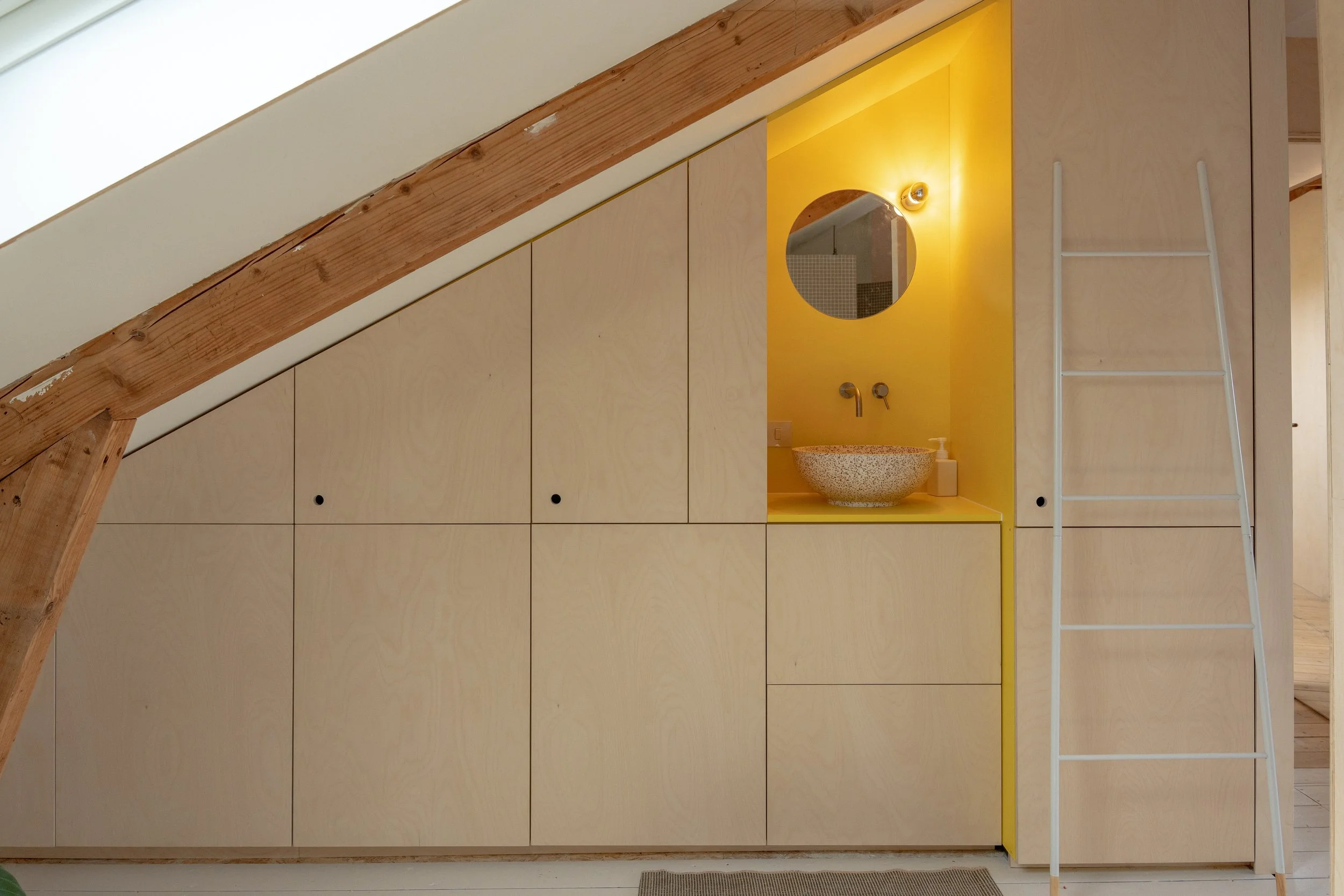 Een wandkast met witte kastdeuren onder een schuine houten plafond. Er is een geel omlijnde nis met een ronde spiegel en een wastafel, evenals een witte ladder naast de nis.