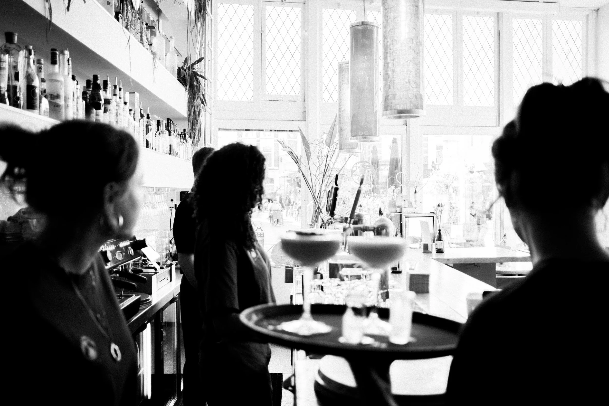 Interieur van een café of restaurant met mensen die achter de bar staan, glazen op een dienblad en grote ramen die fel licht binnen laten.