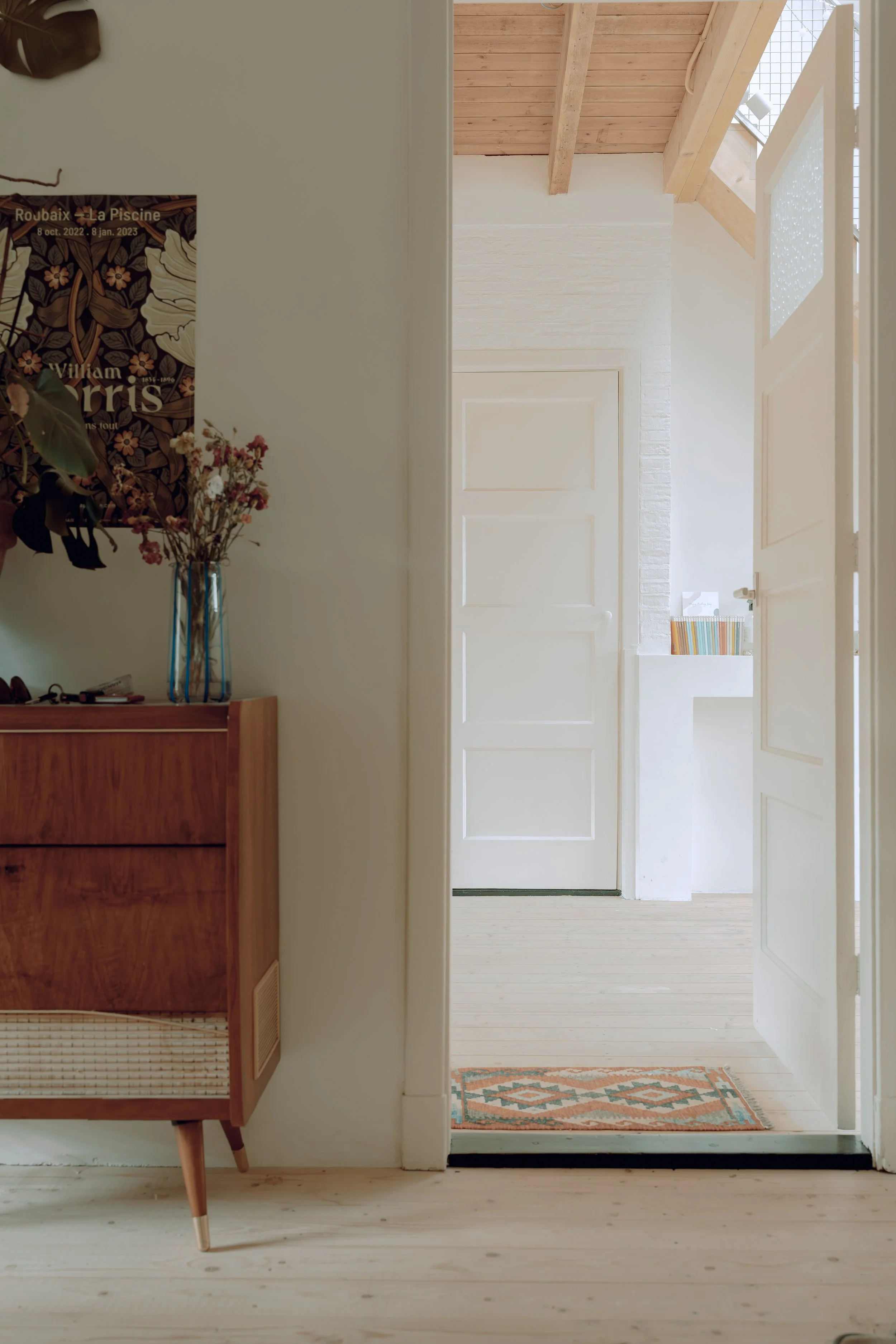 Een woonkamer met een houten vloer en een deur die naar een verlichte hal leidt. Er hangt een poster en er staat een houten kast met bloemen in een vaas.