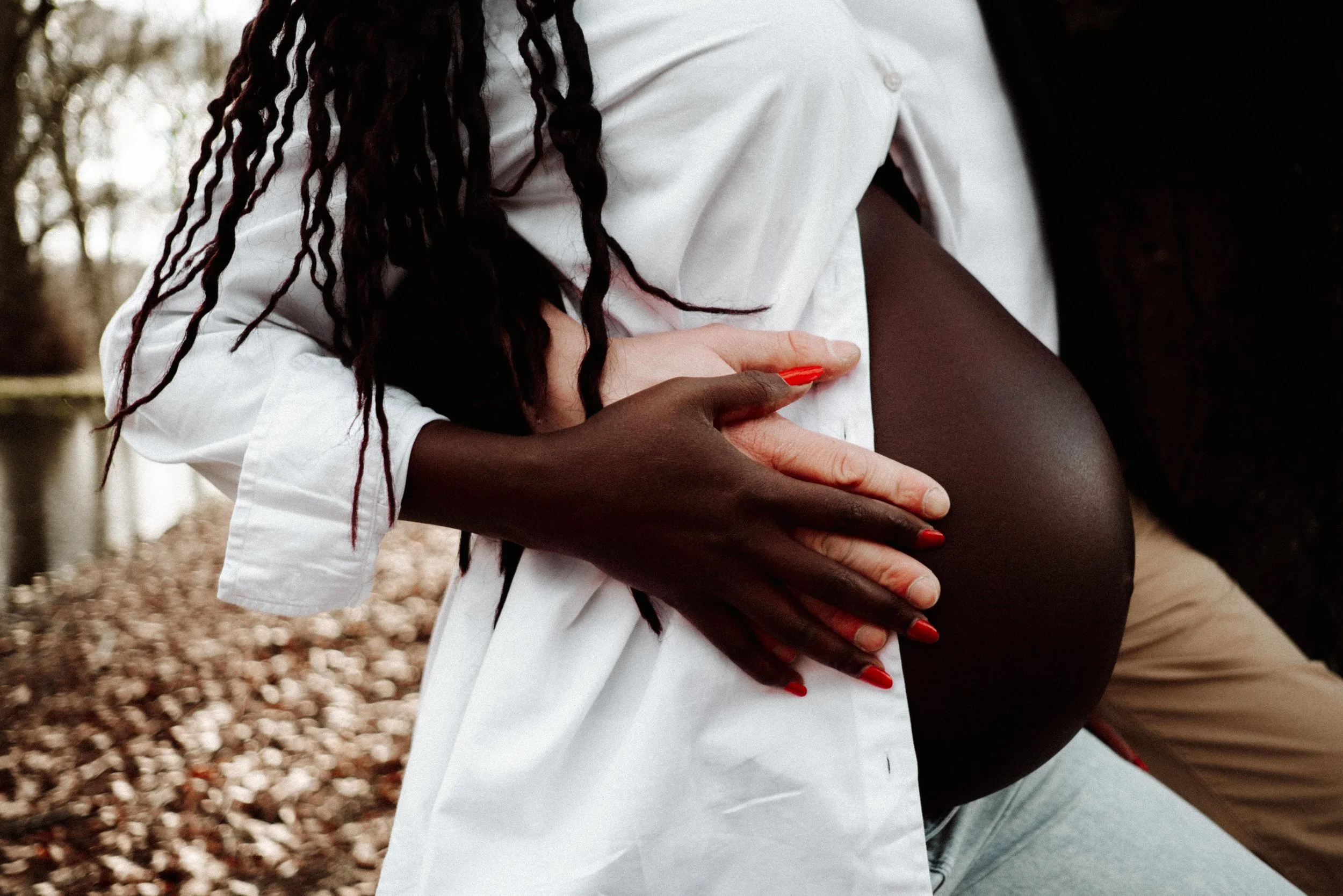 Close-up van een vrouw met donkere huid die haar buik vasthoudt, gekleed in een witte blouse, met lang, gekruld haar, op een buitenlocatie met bladeren op de grond. zwanger koppel stelletje ouders baby zwangerschap fotografie Arnhem 