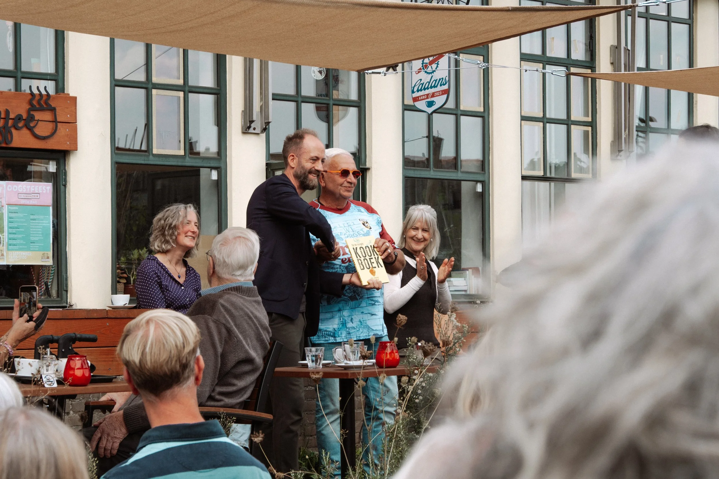 Een groep mensen bij een buitenbijeenkomst, met één persoon die een boek vasthoudt dat 'KEUKENBOEK' heet, terwijl anderen applaudisseren. Er hangt een bord met de naam 'Sadans' en er zijn tafels en stoelen met decoraties.