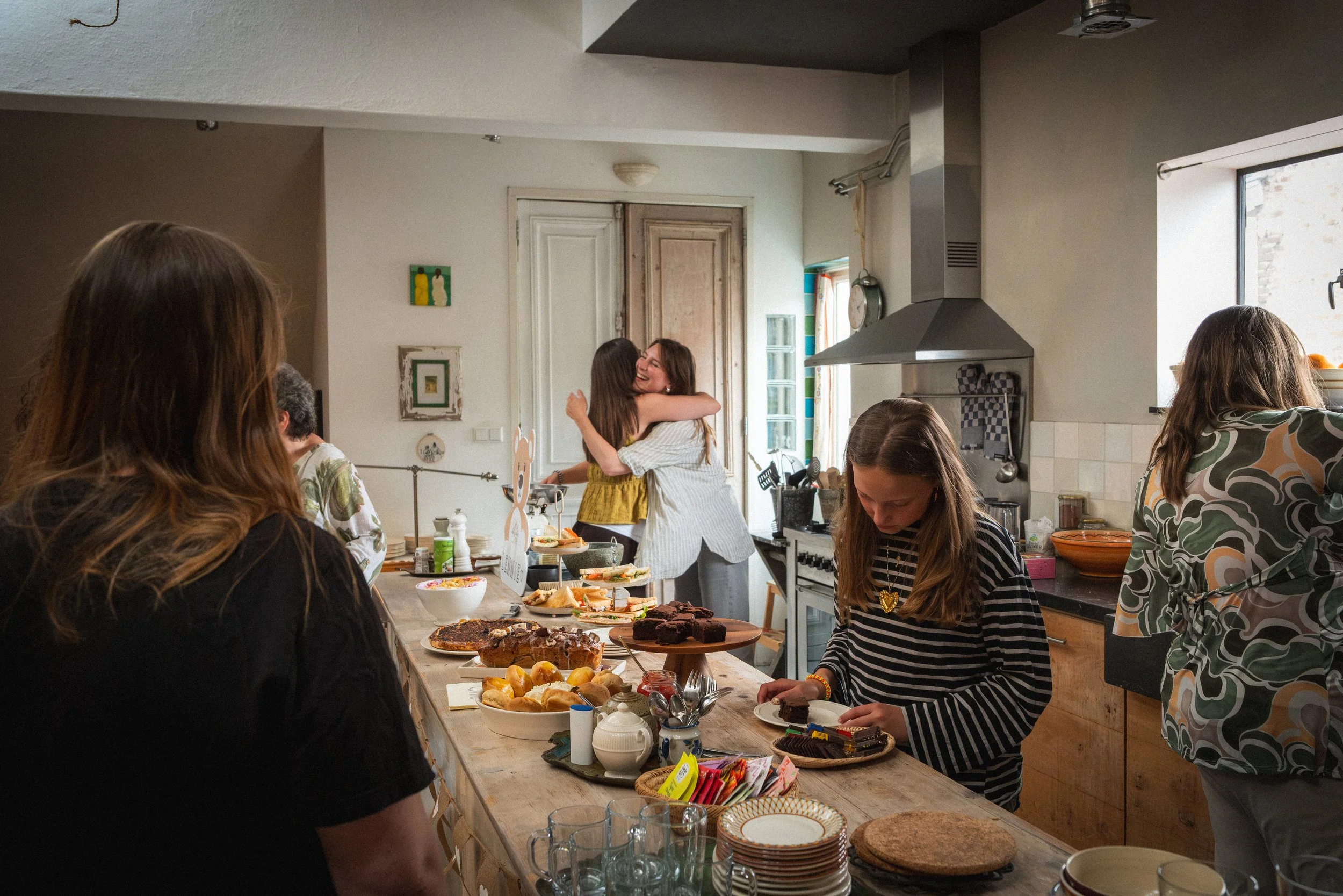 Mensen in een keuken vieren een feest met veel eten en drinken, mensen knuffelen en glimlachen, vooral twee vrouwen die elkaar omarmen.