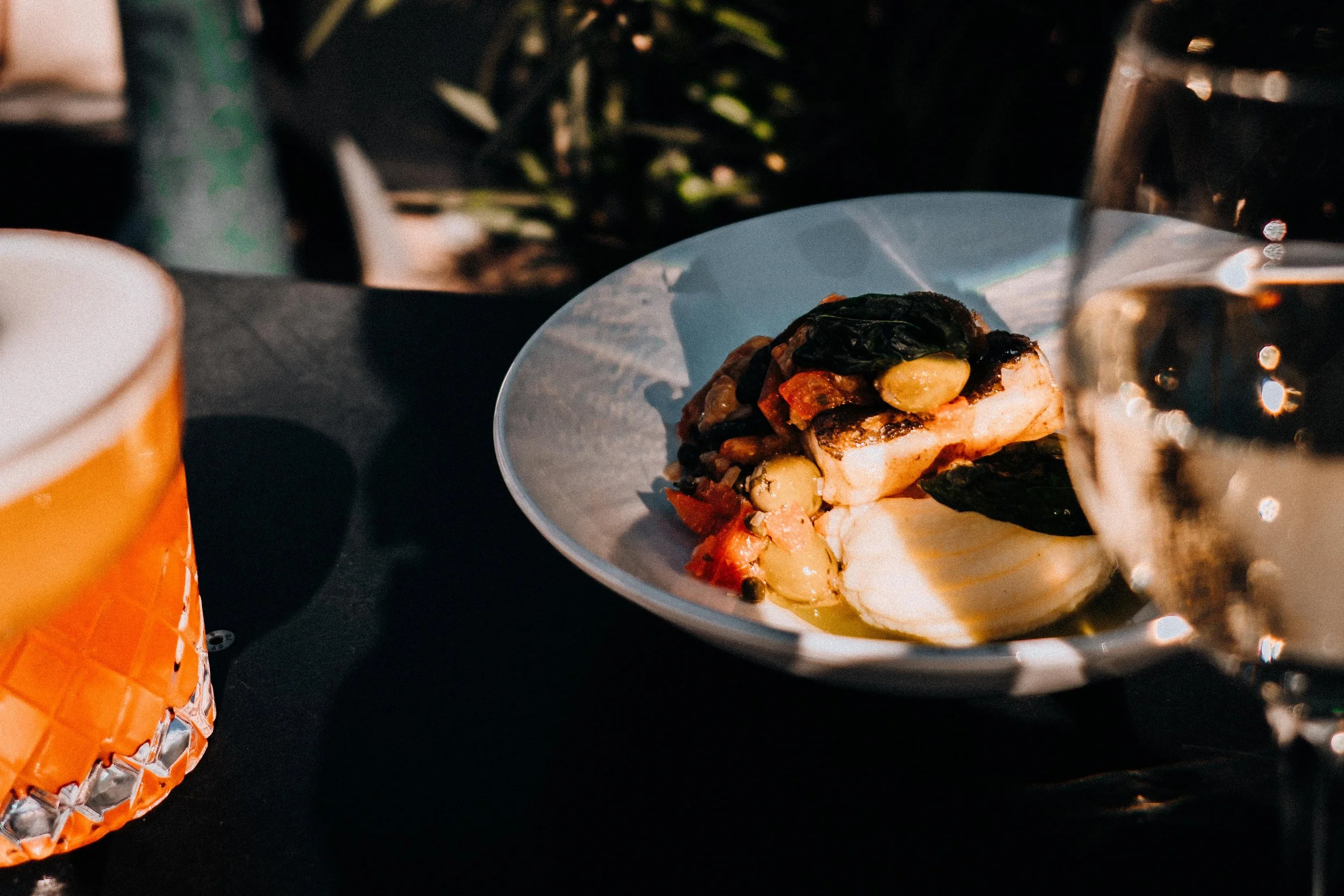 Een gerecht met gegrilde vis, spinazie, knoflook en tomaat op een wit bord, omringd door een glas bier, een blikje en een oranje beker.