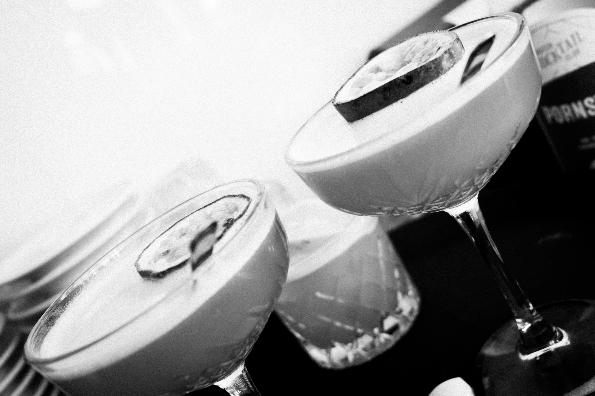 Twaalf cocktails in martiniglaasjes, afgedekt met een schijfje citroen, op een witte plaat, zwart-wit foto.