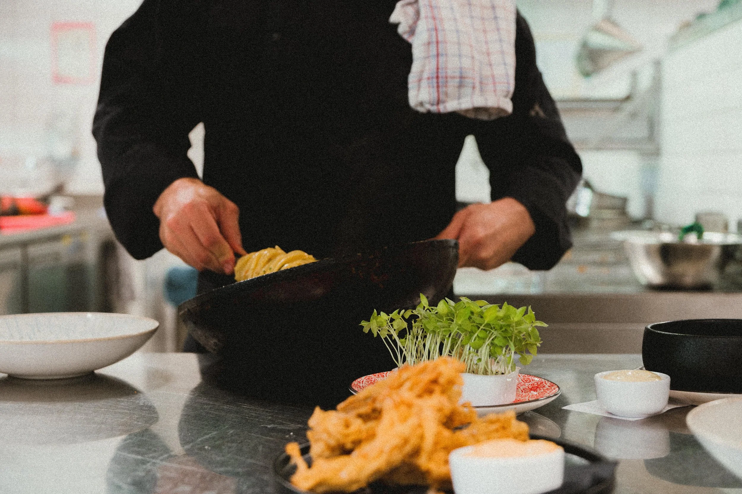 Persoon bereidt voedsel in een professionele keuken, met verschillende gerechten en ingrediënten op de voorgrond.