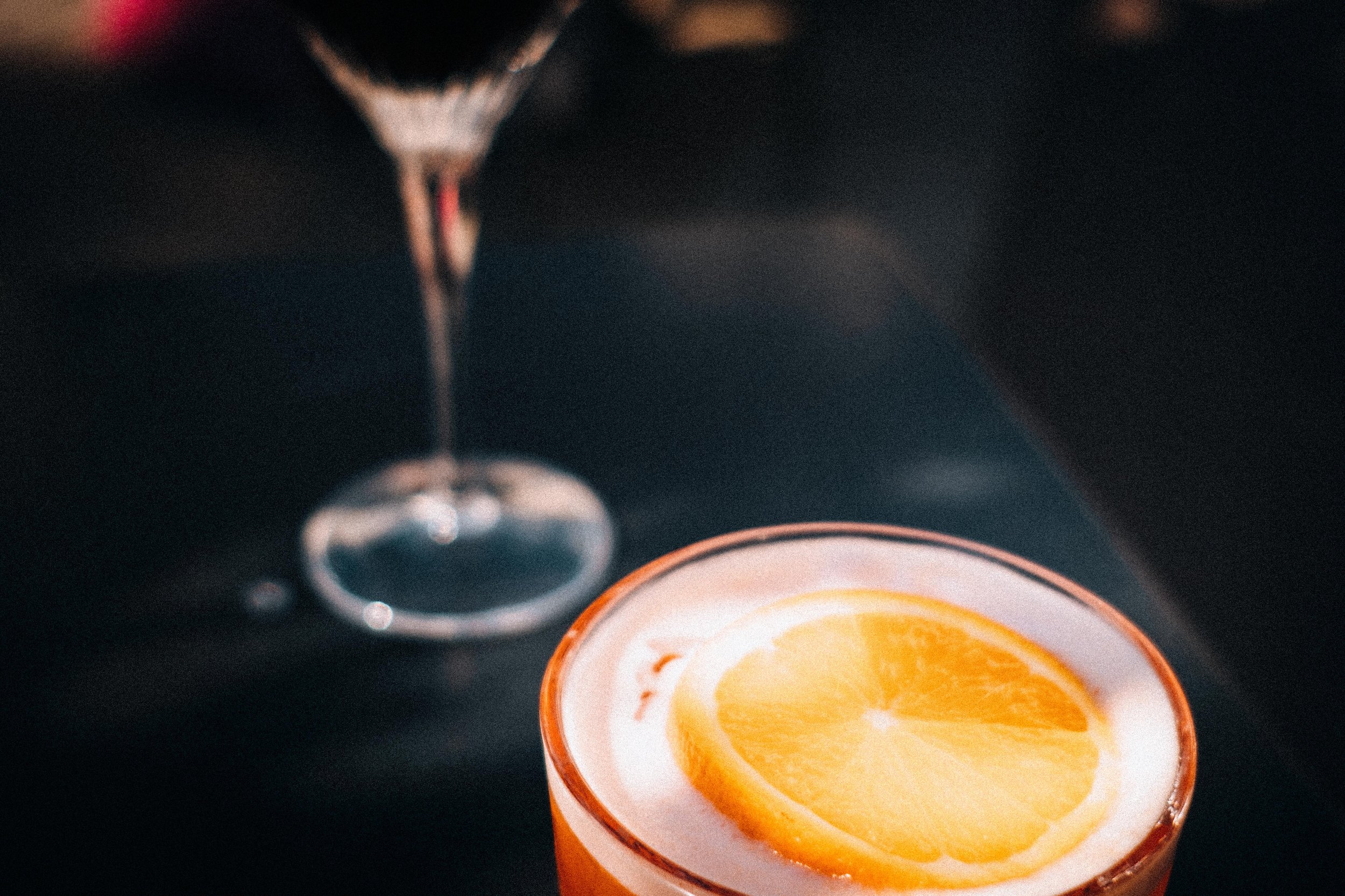 Close-up van een brandende kaars in een oranje houder, met een glas en een andere kaars op de achtergrond.